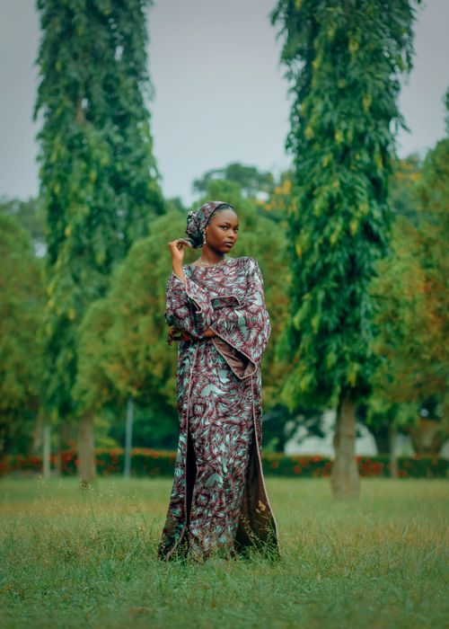 Free A woman in a patterned dress poses in a park, surrounded by tall trees, capturing grace and nature's beauty. Stock Photo