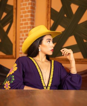 Stylish woman in a purple embroidered dress and yellow hat seated indoors, radiating elegance and modern fashion.