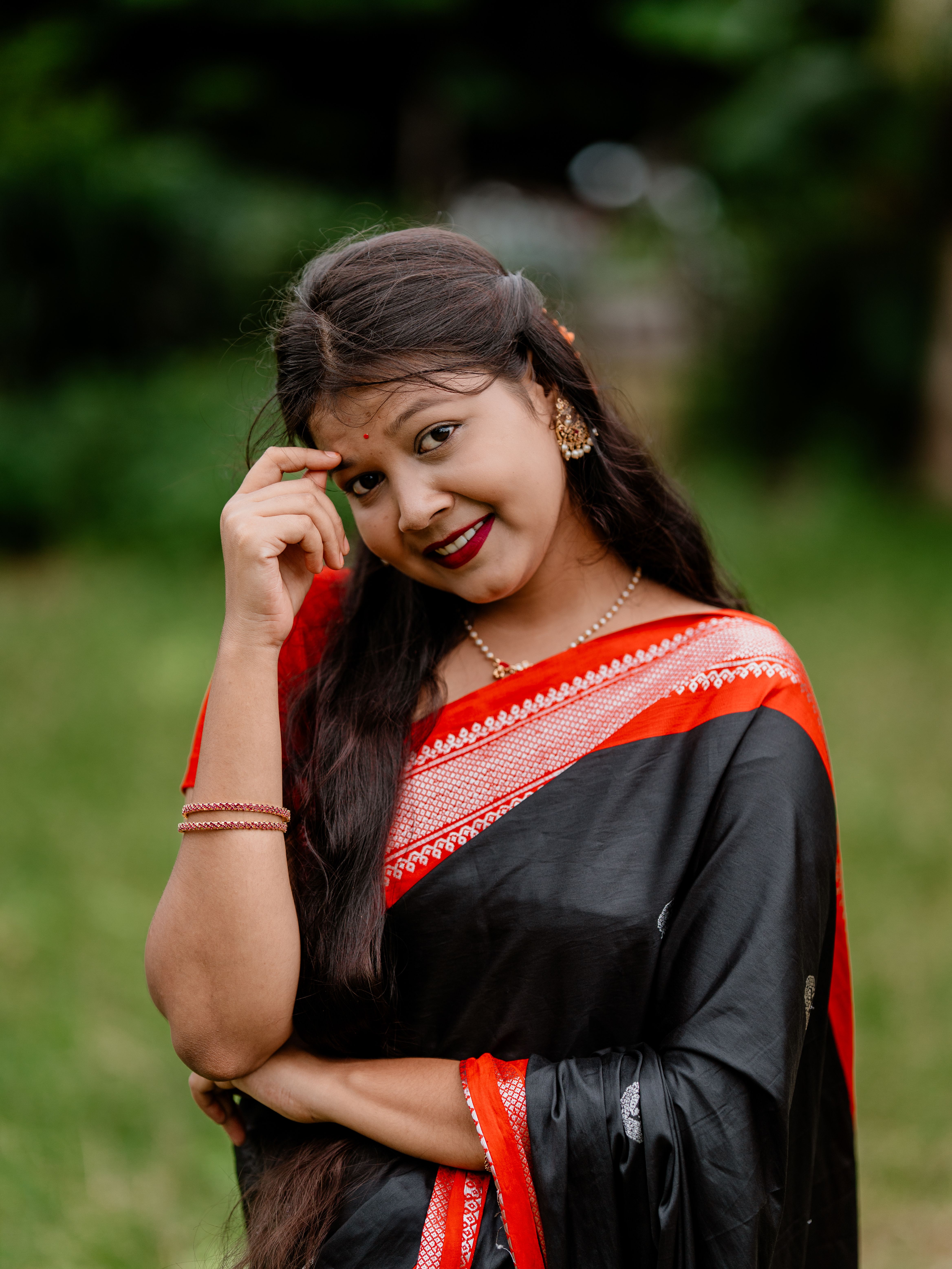 Portrait of a smiling woman in a traditional black and red sari in a lush green outdoor setting.