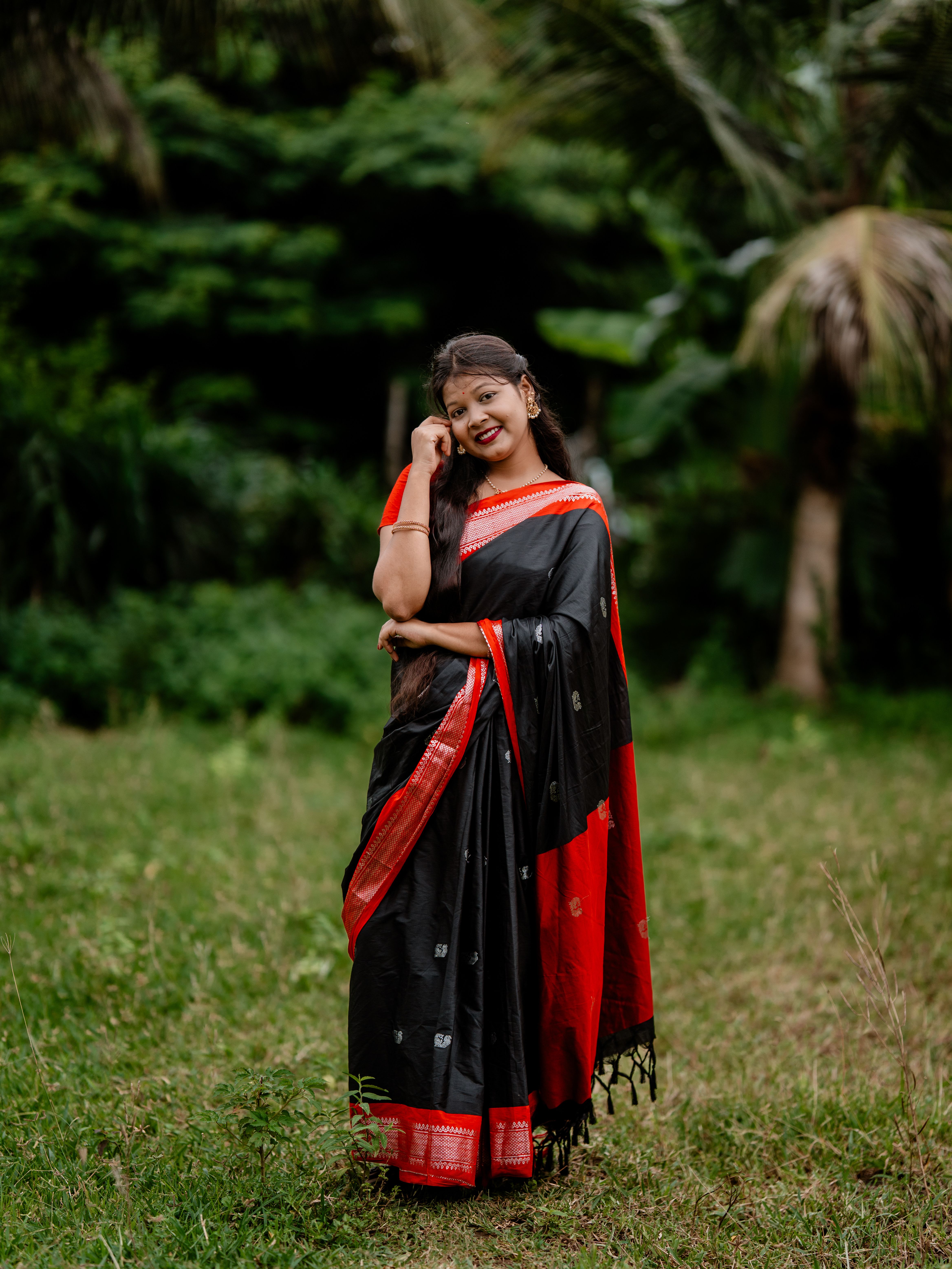 A woman in a red and black saree smiling gracefully amidst lush greenery in daylight.