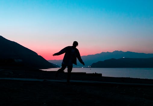 Silhouette of man balancing on one leg on peaceful hillside near calm river in sunset time