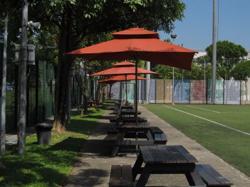 Free Picnic tables with red umbrellas beside an outdoor sports field under a clear blue sky. Stock Photo