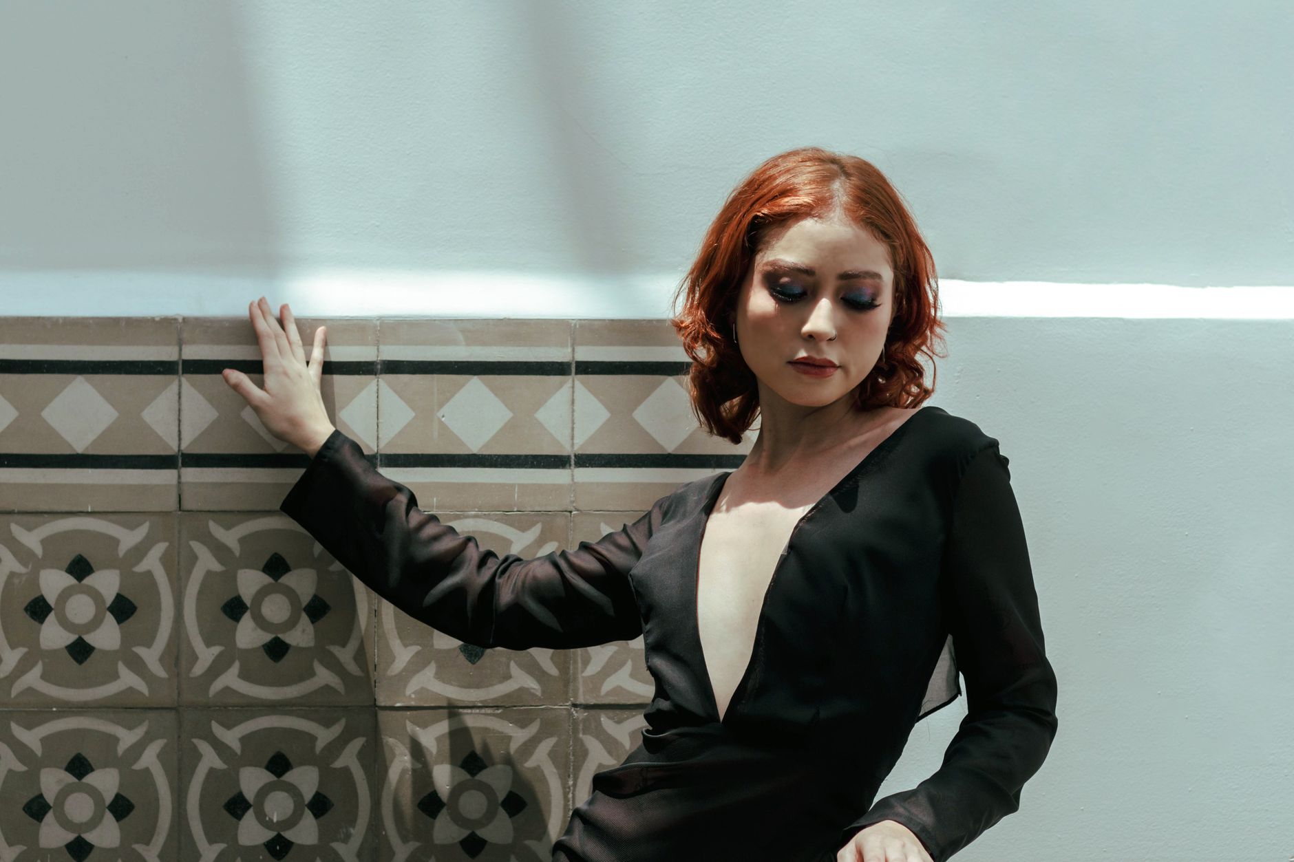 Stylish woman in a black dress posing against a decorative wall under natural lighting.