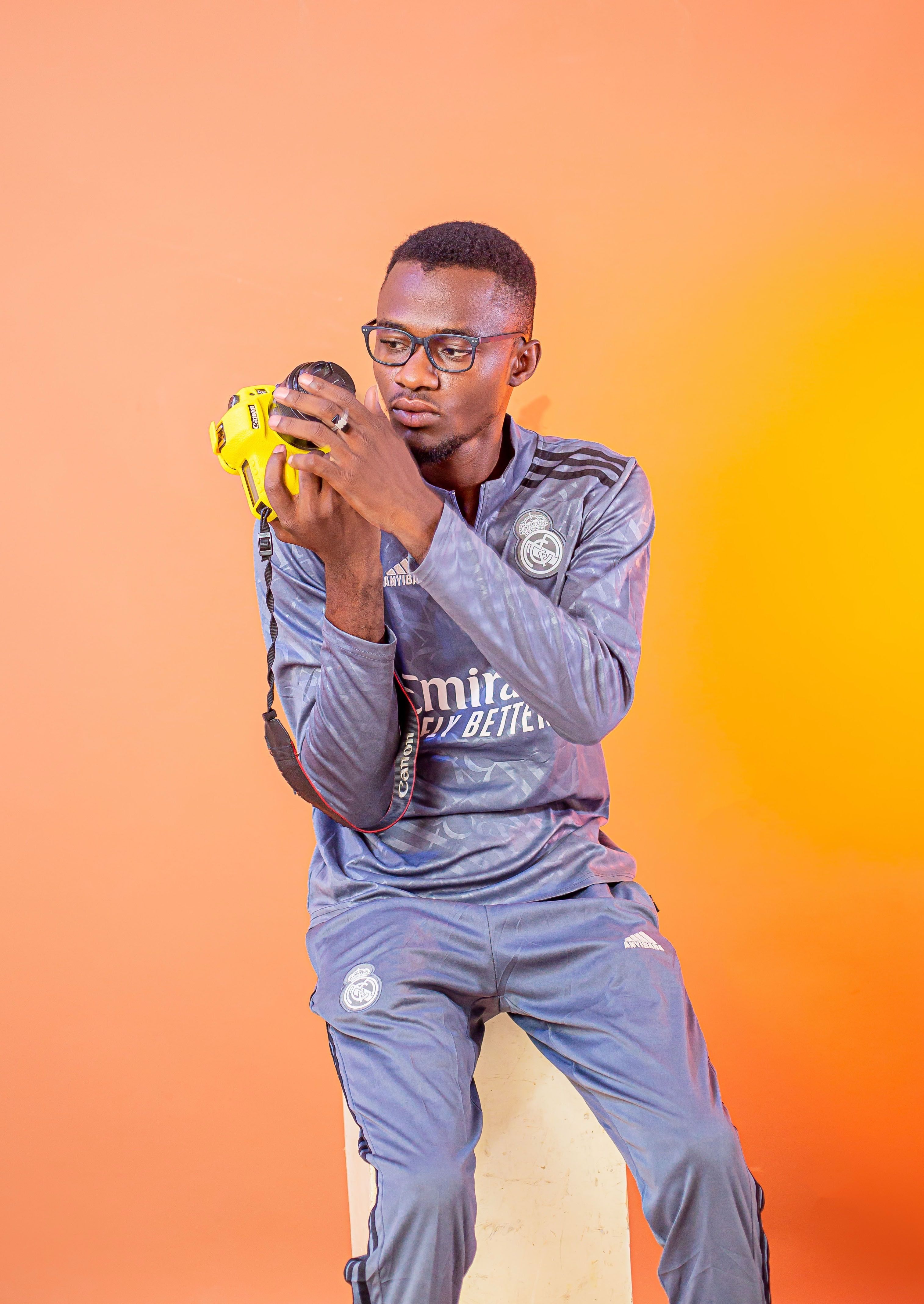 Young man in sportswear holding a yellow camera against an orange backdrop.