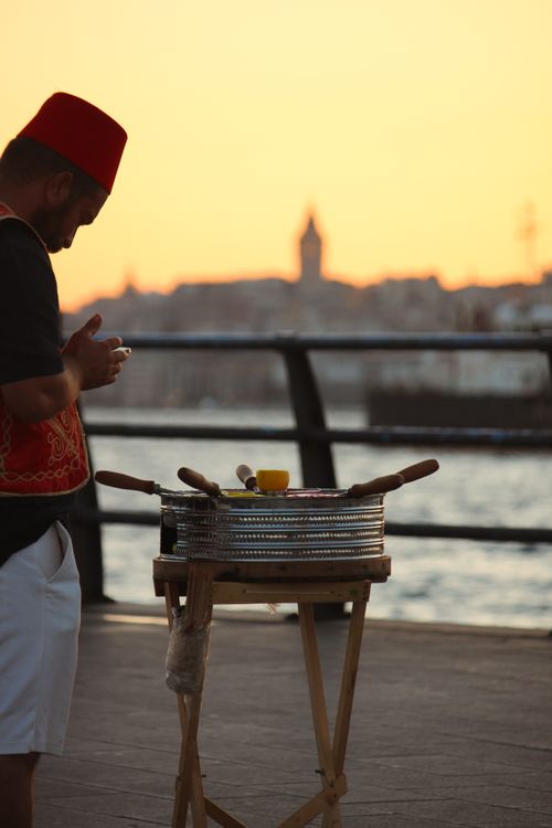 Free A street vendor wearing a fez prepares food against a scenic sunset in Istanbul with a historic tower silhouette in the background. Stock Photo