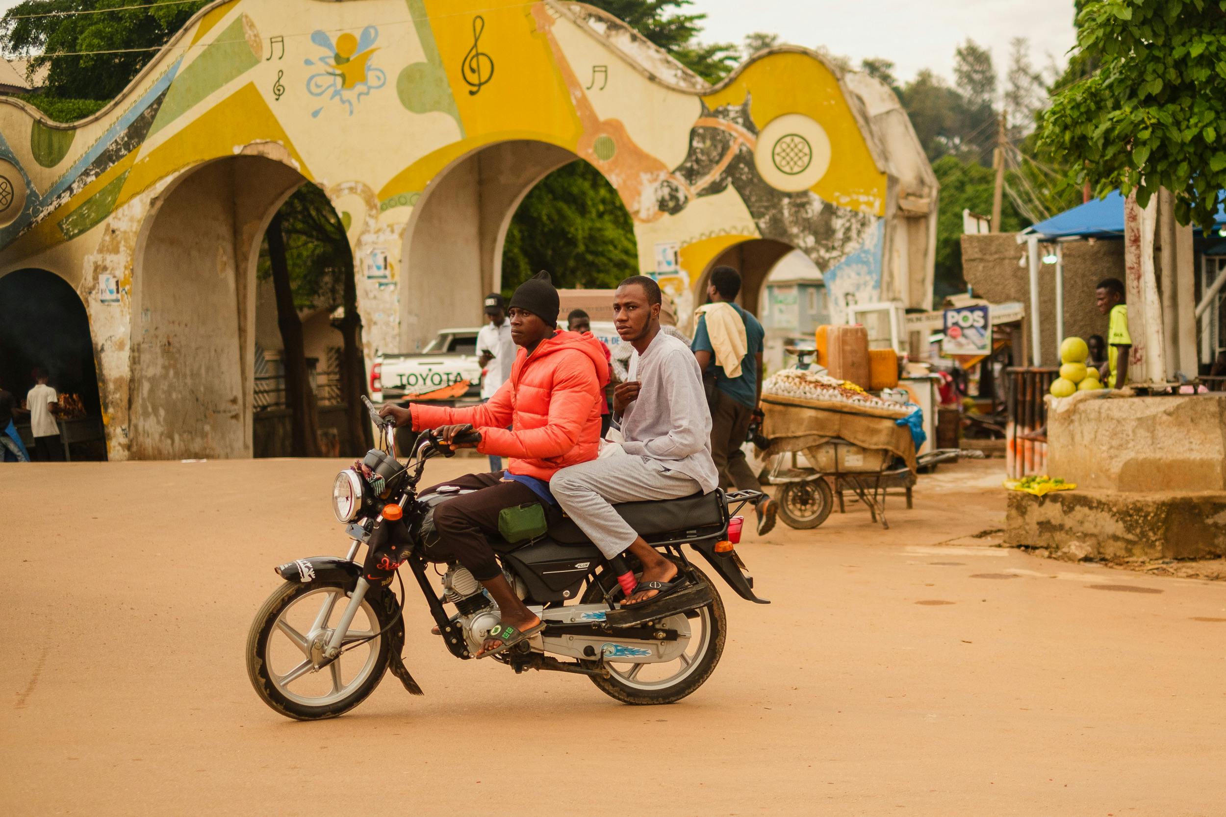 Two men on a motorbike passing through a vibrant street in Kaduna, Nigeria.