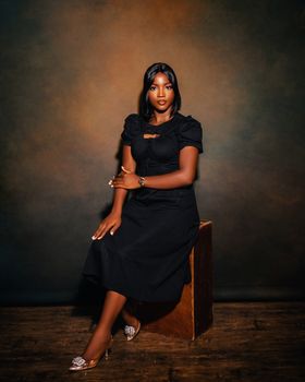 Elegant woman posing in a black dress against a dark background, seated gracefully.