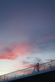 A lone person walking along a bridge railing under a colorful sunset sky.