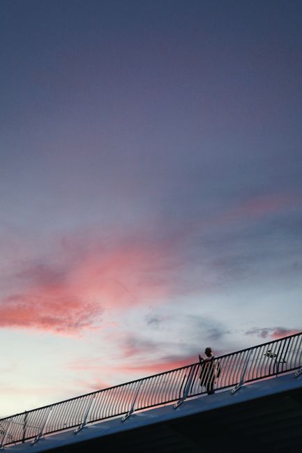 A lone person walking along a bridge railing under a colorful sunset sky.