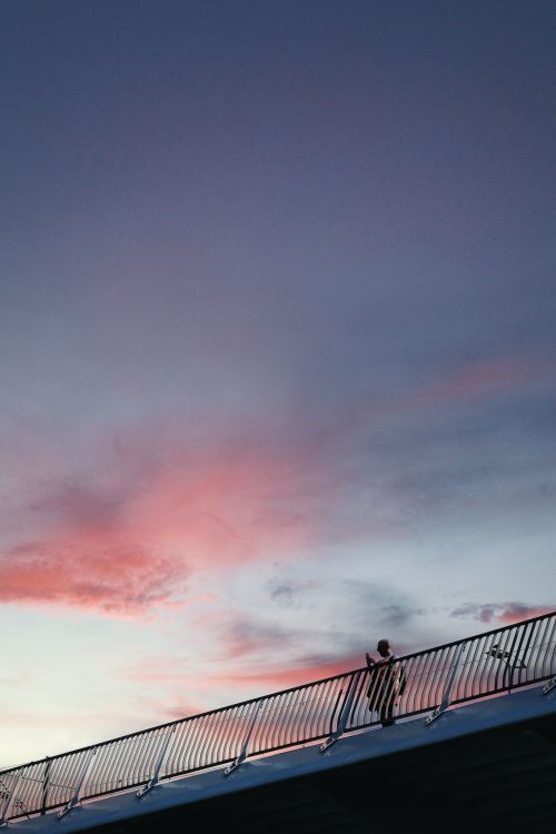 Free A lone person walking along a bridge railing under a colorful sunset sky. Stock Photo
