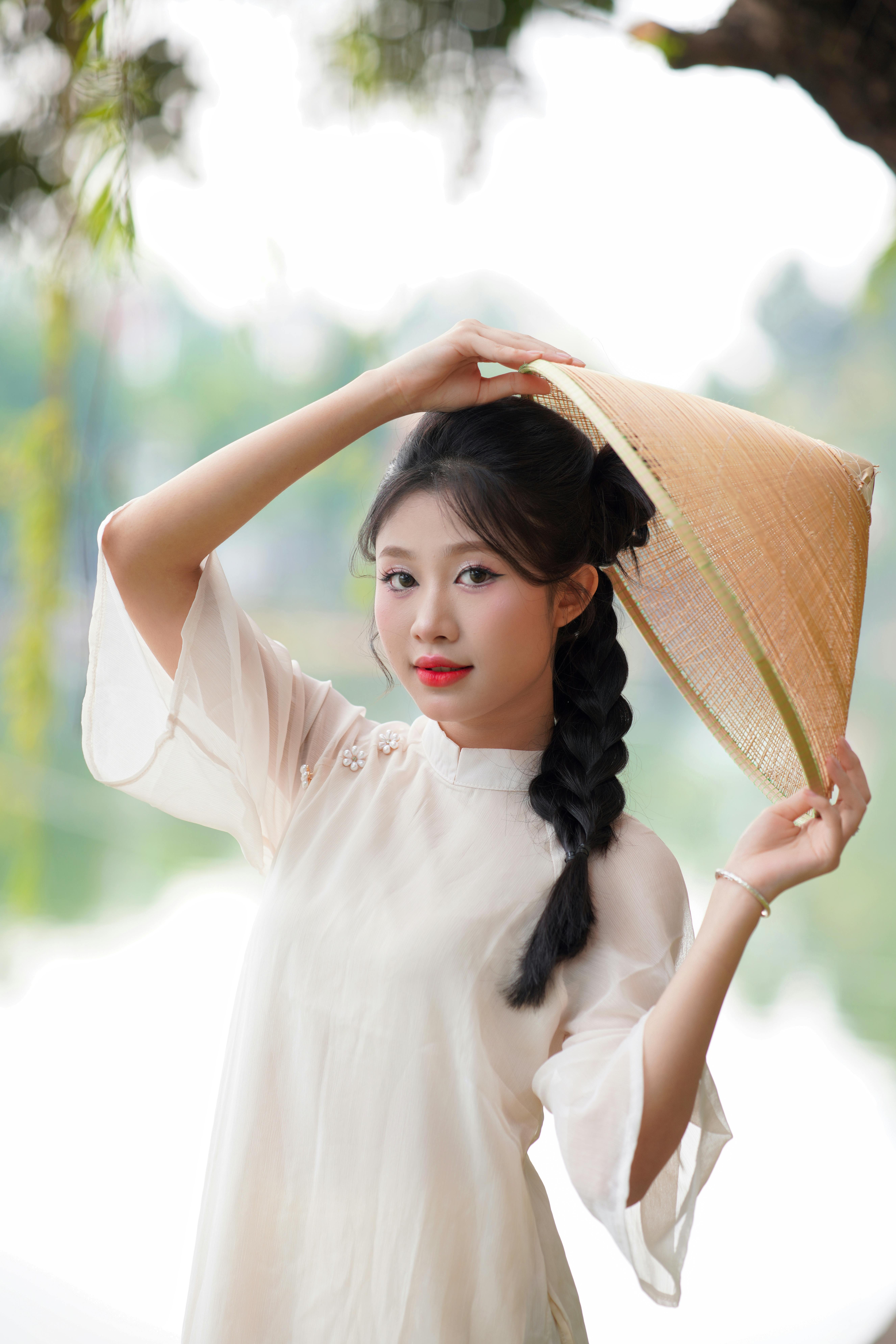 Elegant portrait of a young woman wearing a conical Asian hat in a serene outdoor setting.