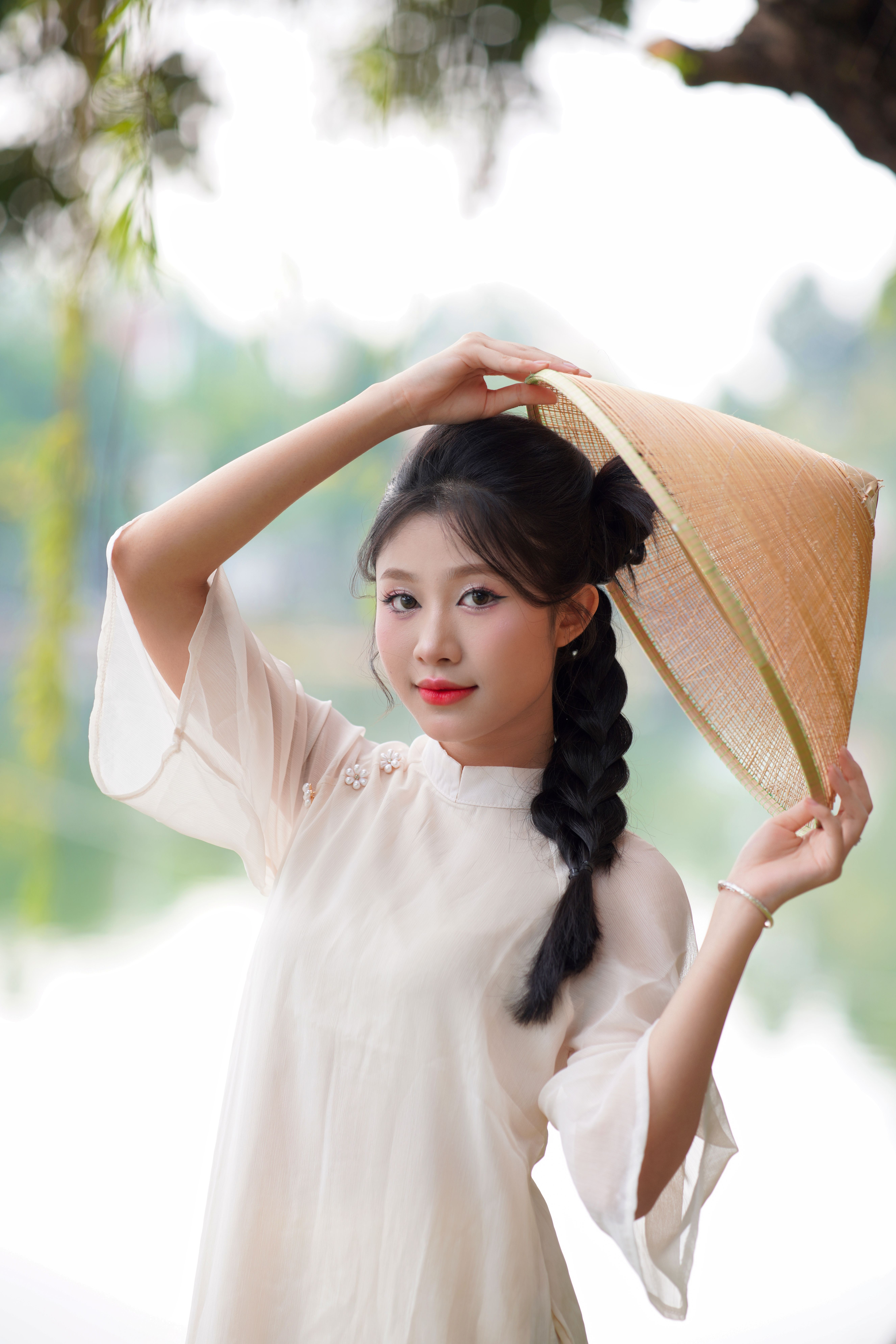 Portrait of a young woman in traditional Asian attire holding a conical hat outdoors.