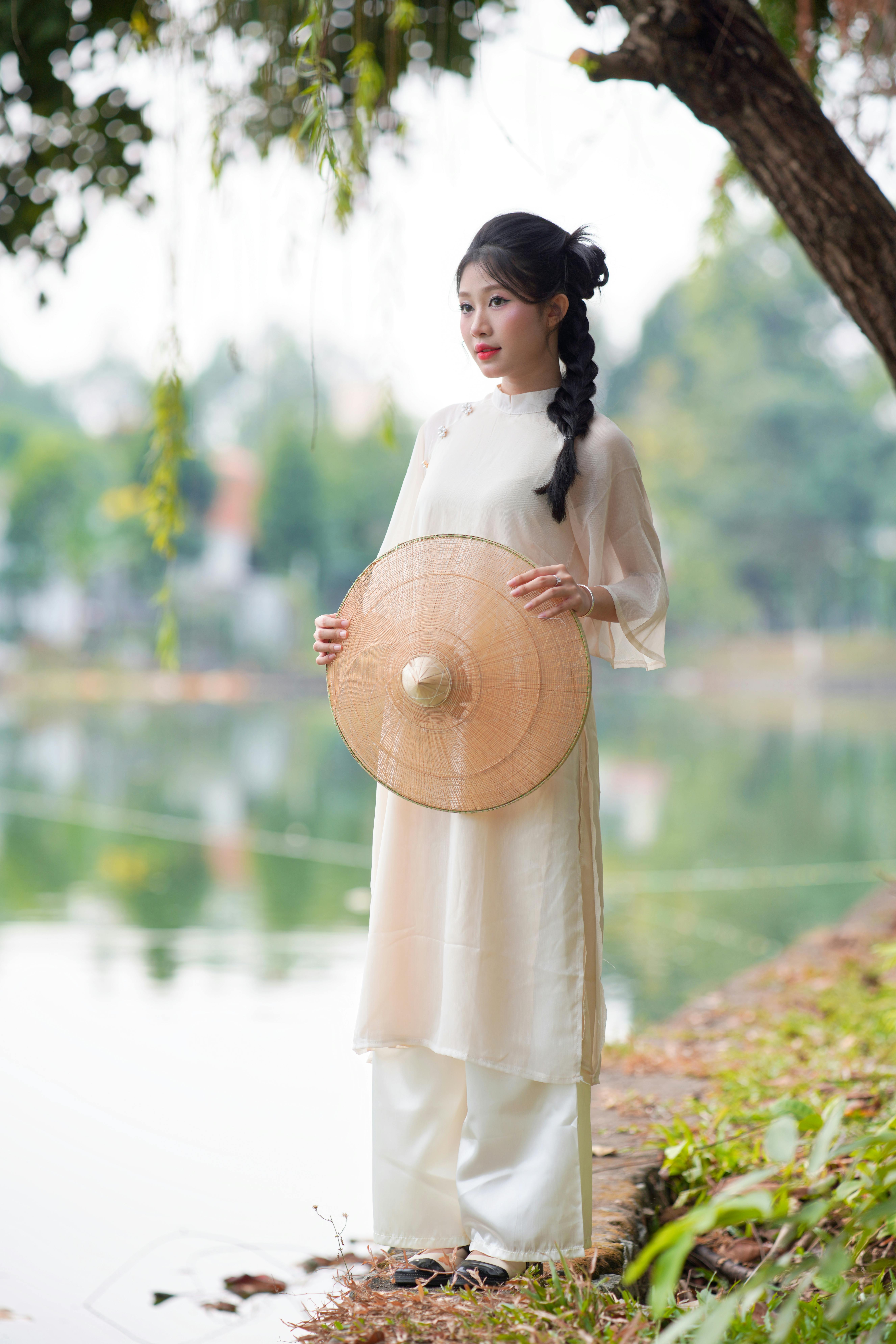 Woman in serene outdoor setting holding a conical hat, symbol of elegance.