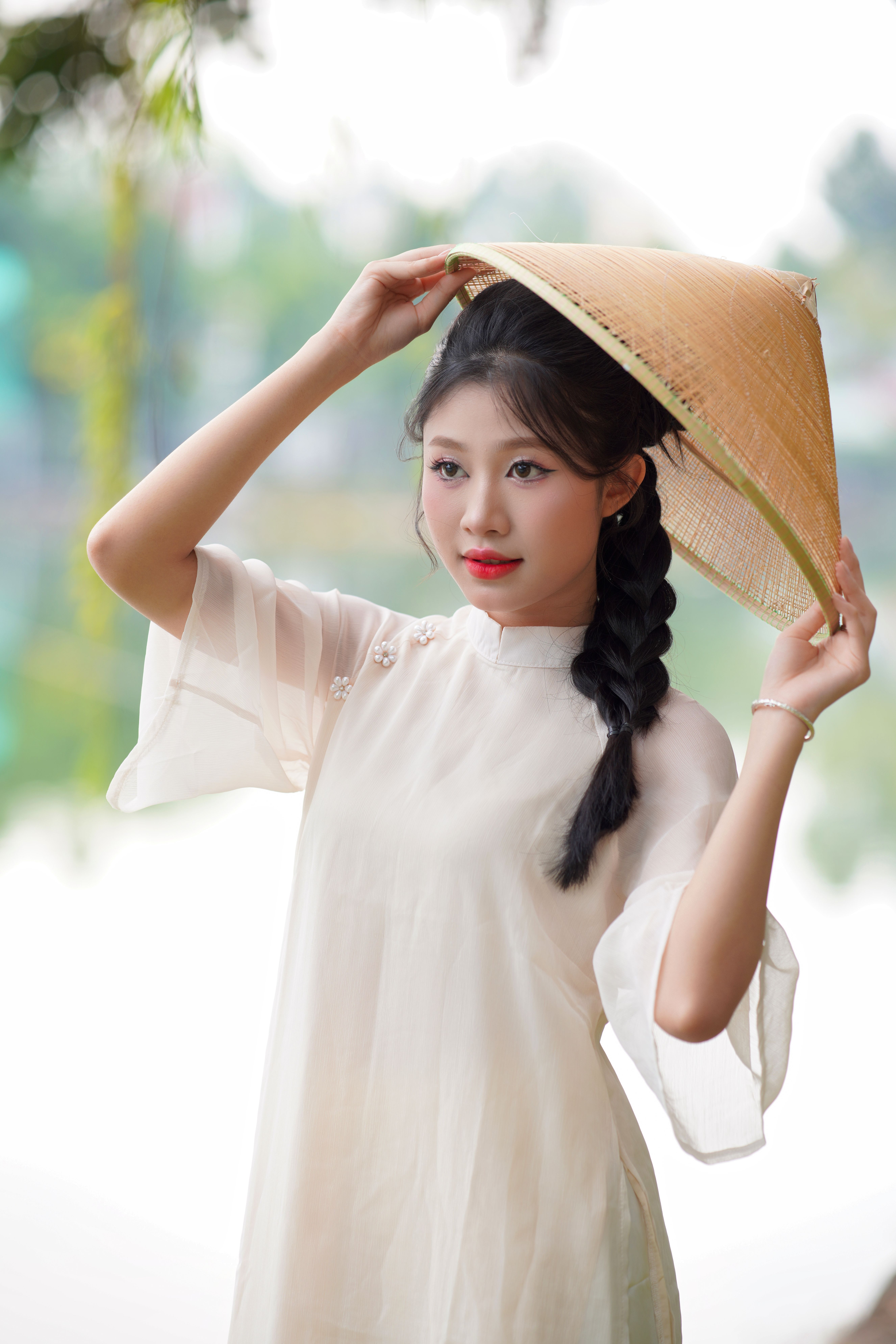 A young woman wearing an Ao Dai and conical hat, standing outdoors with a serene expression.