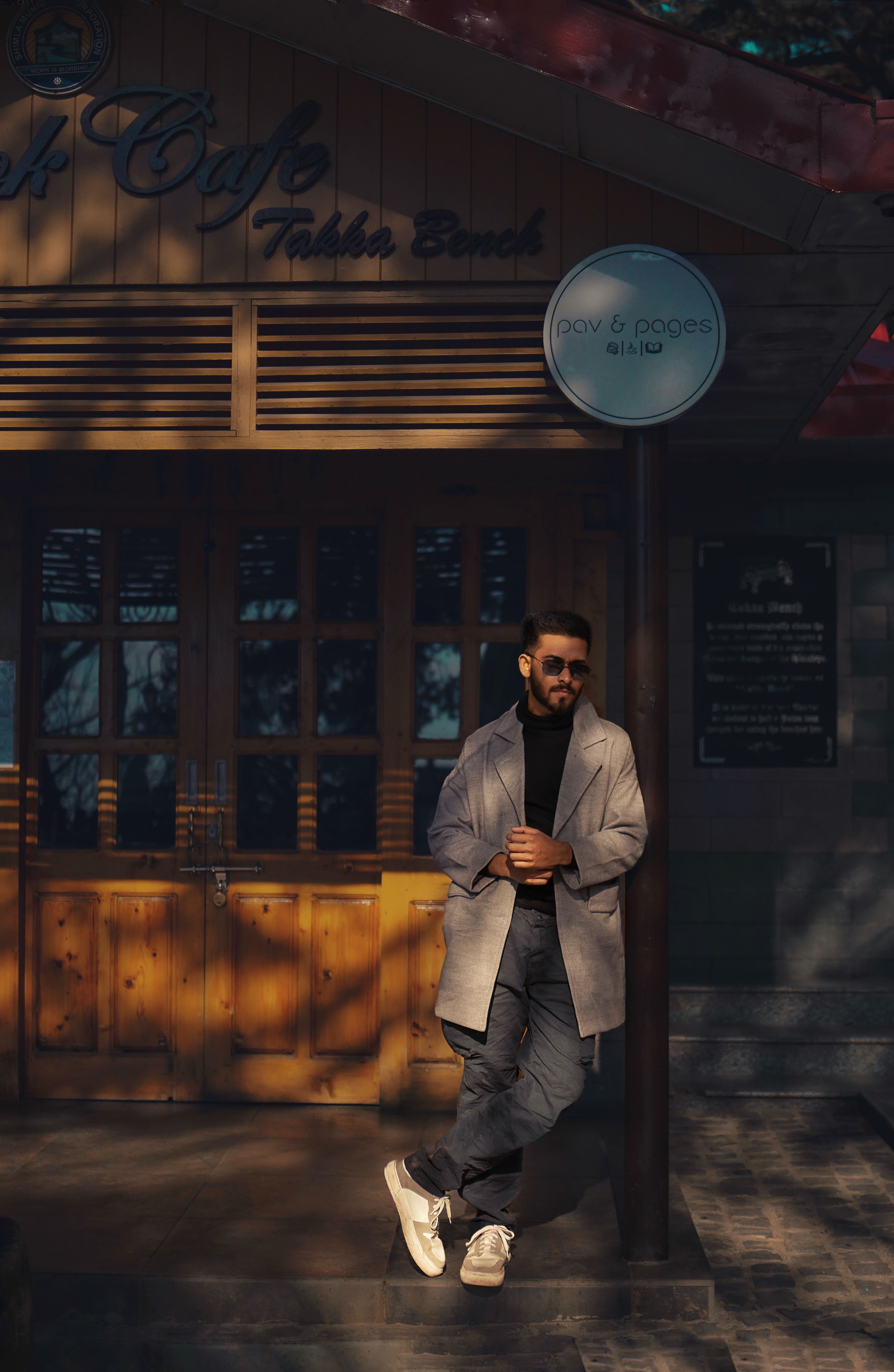 Man in coat and sunglasses leans against a cafe in Shimla, India, on a sunny winter day.