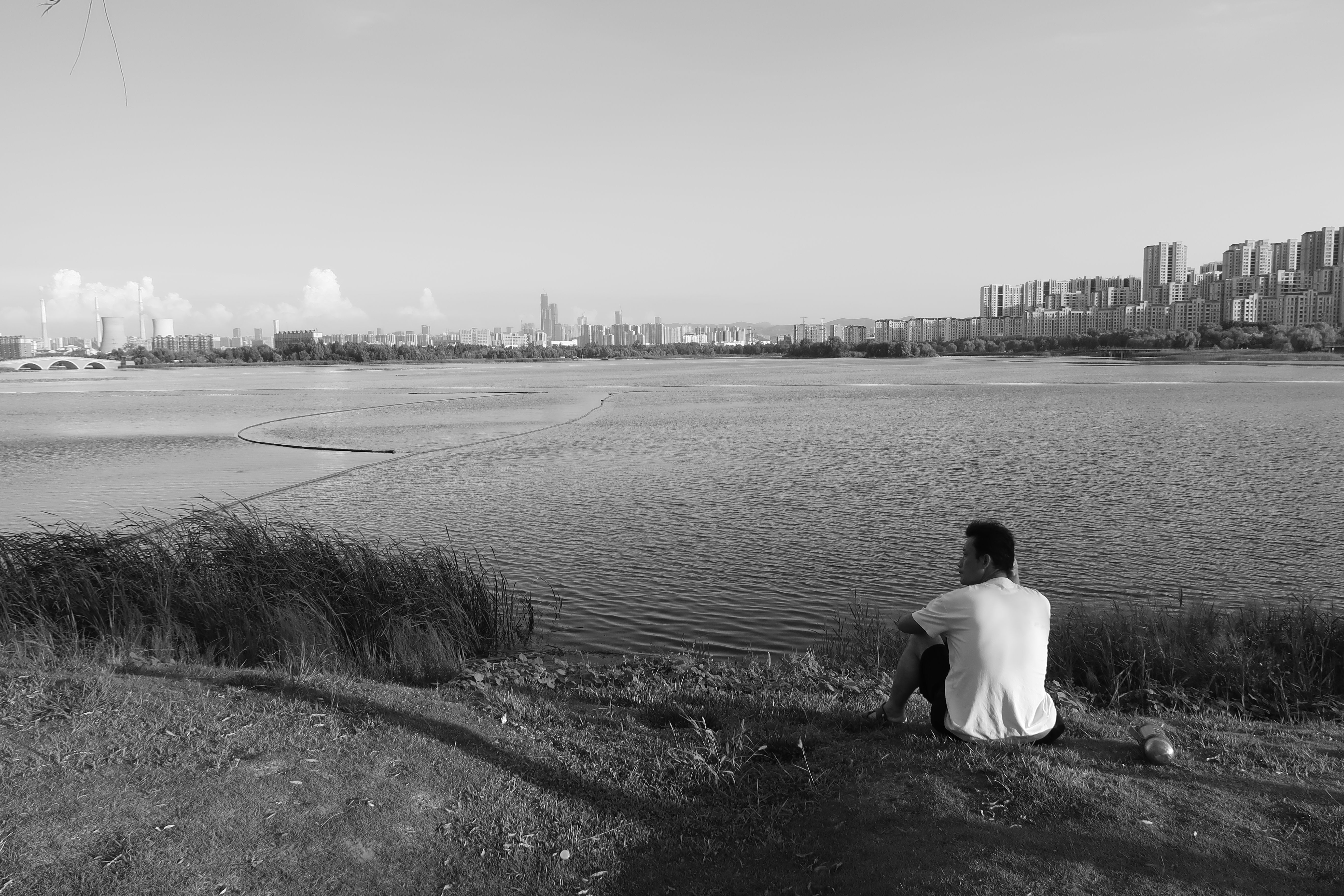 A lone man sits by a river, observing a city skyline in monochrome.