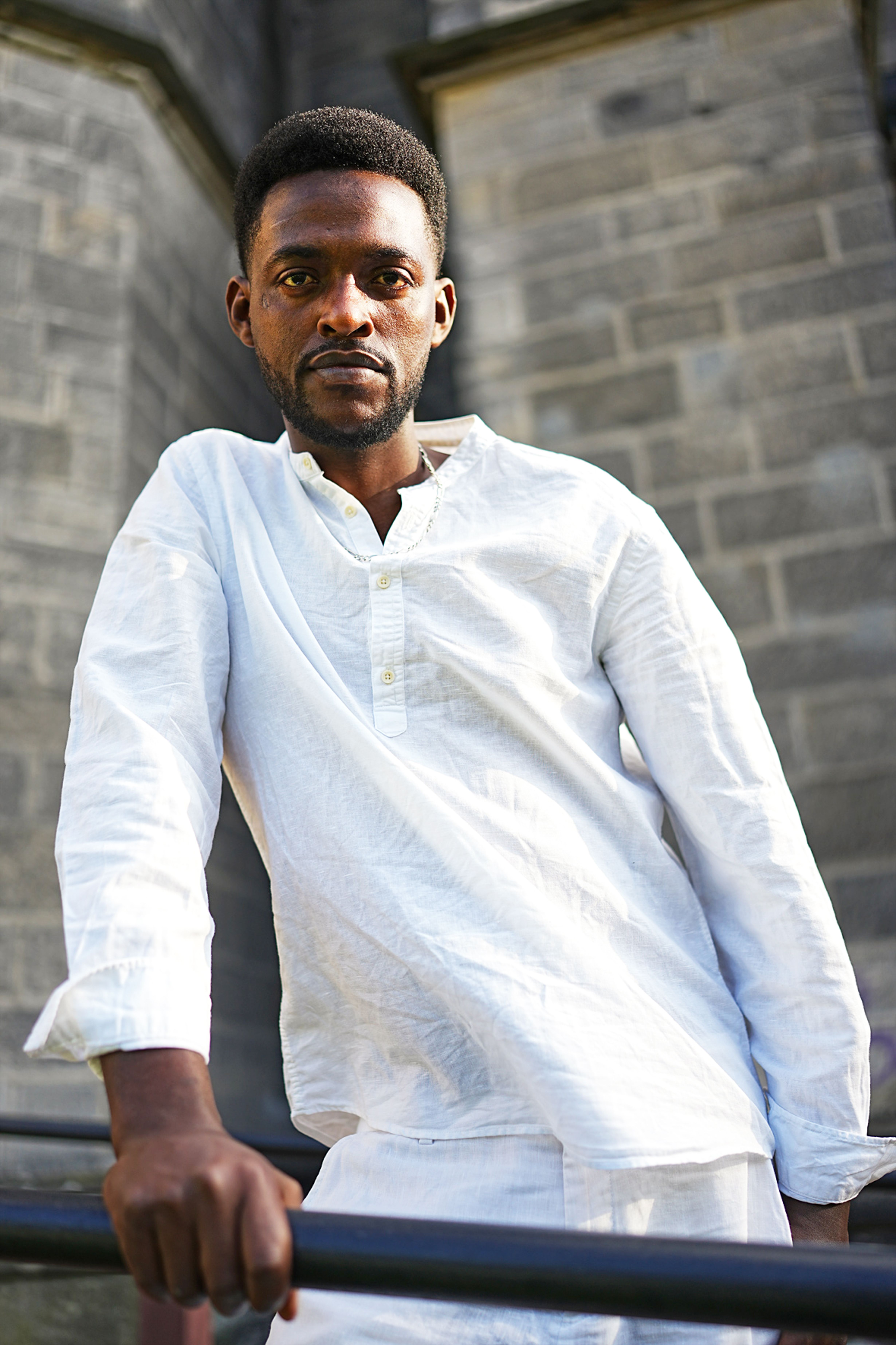 An adult man in a white shirt poses outdoors against a stone wall, conveying confidence.