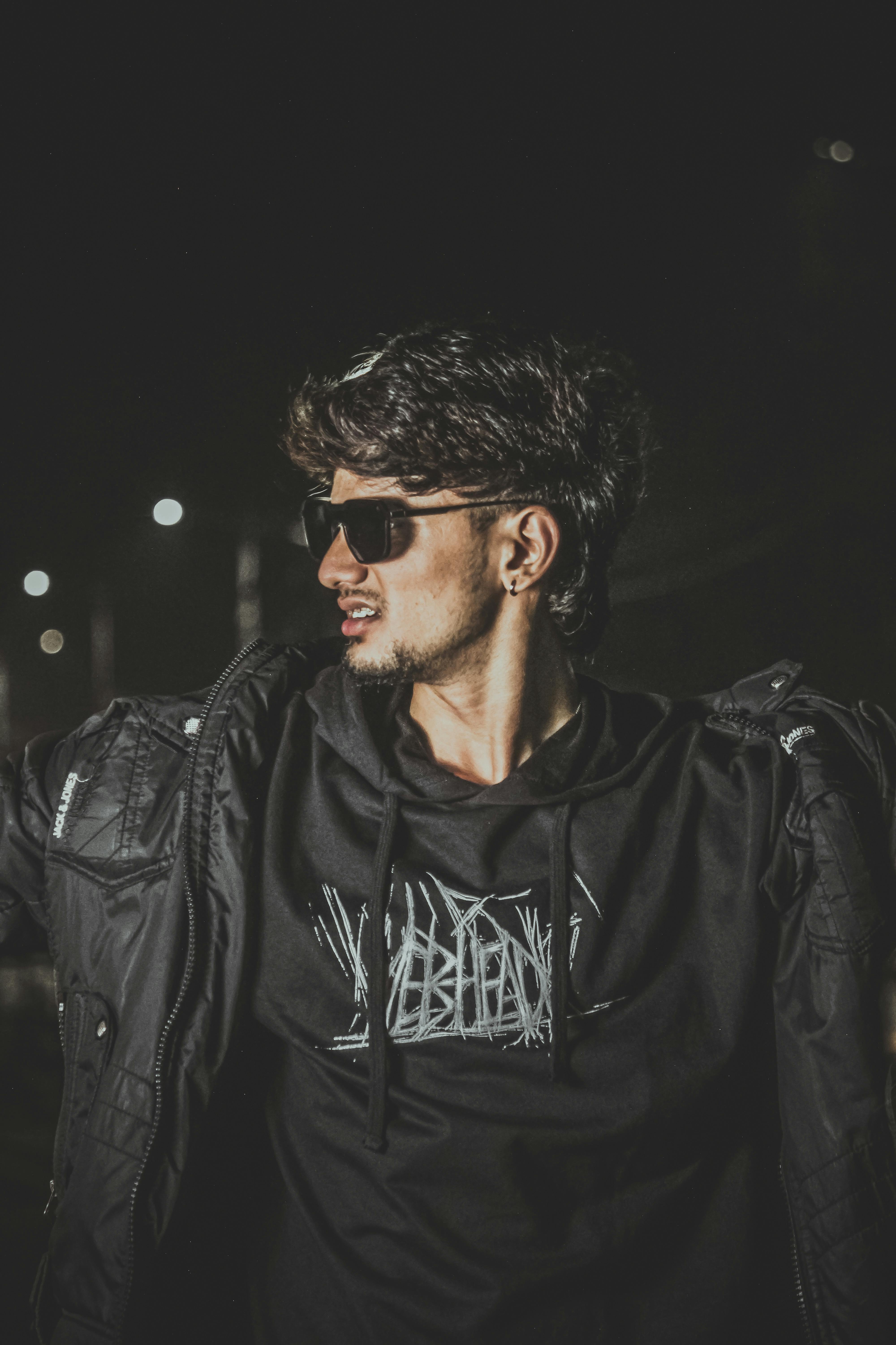 Fashionable young man wearing sunglasses and dark clothing, captured in a moody nighttime setting.