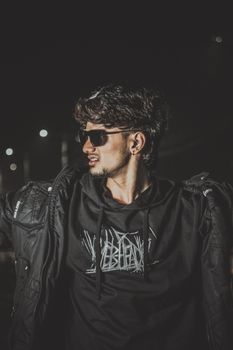 Fashionable young man wearing sunglasses and dark clothing, captured in a moody nighttime setting.