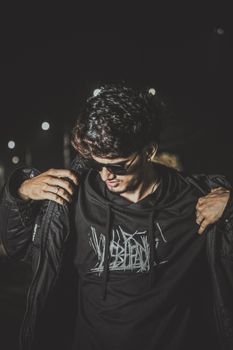 Fashionable man in hoodie and jacket, sunglasses on, posing at night with city lights.