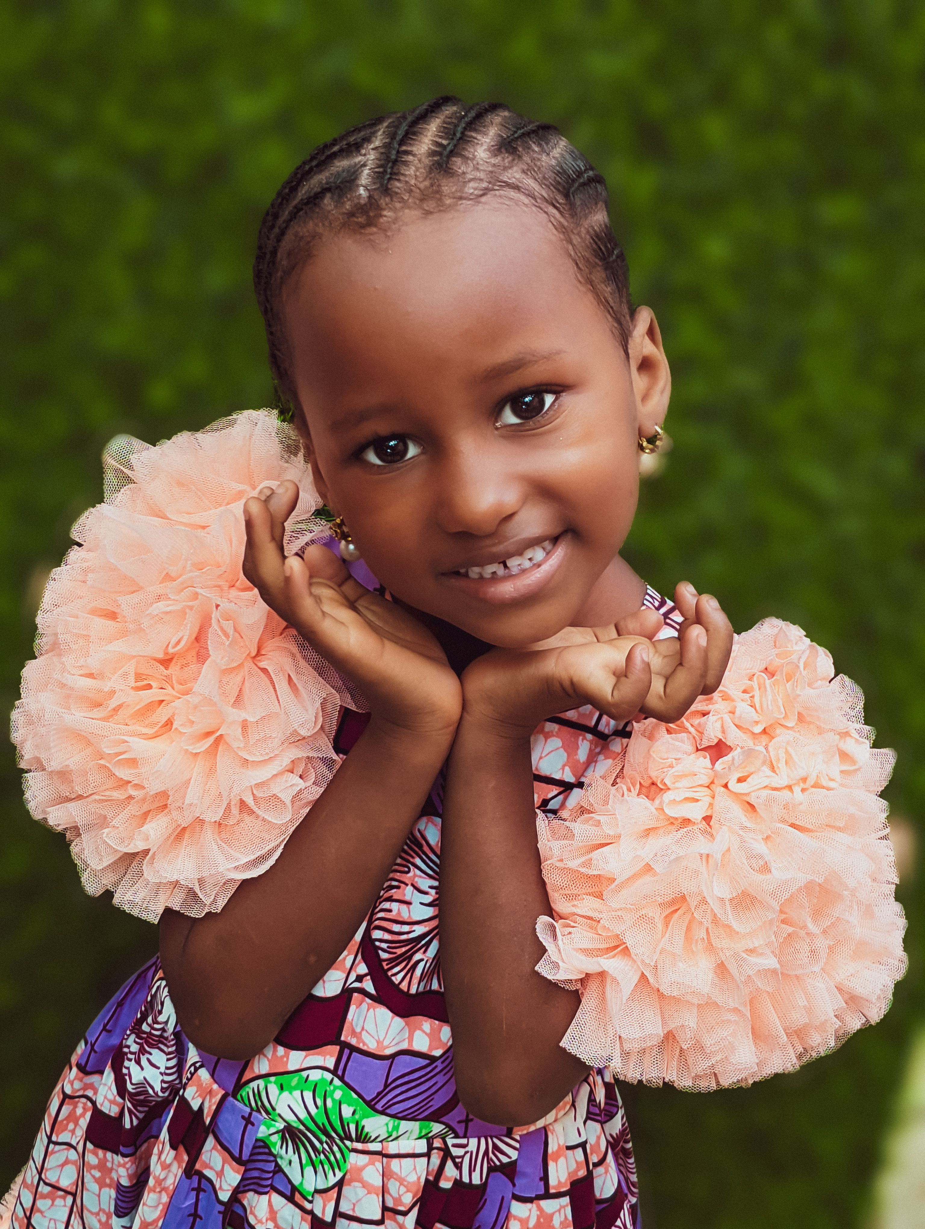 Smiling African girl in colorful traditional attire, posing outdoors with vibrant ruffled sleeves.
