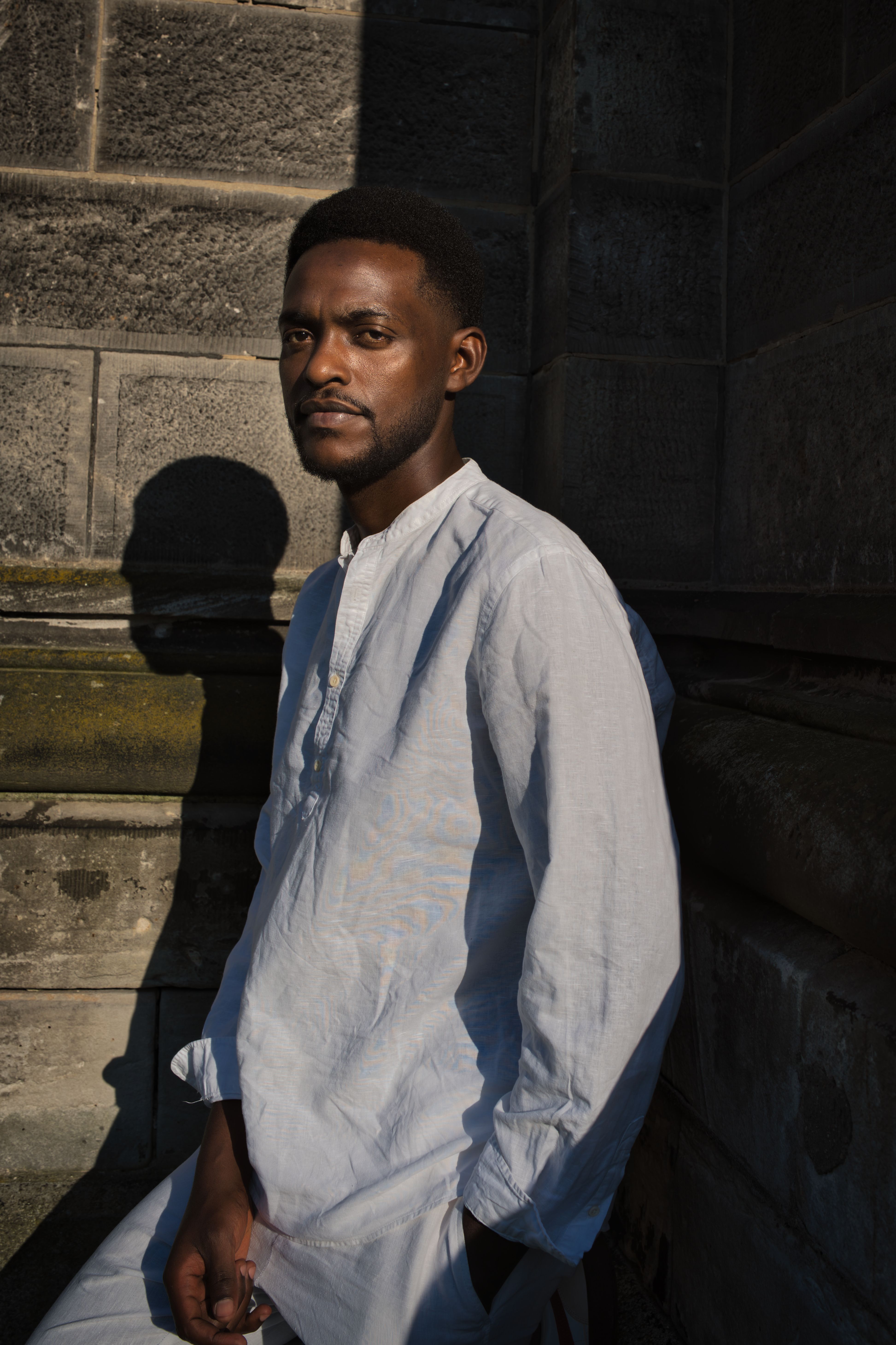 Man in a white shirt leans against a stone wall, highlighted by warm sunlight.
