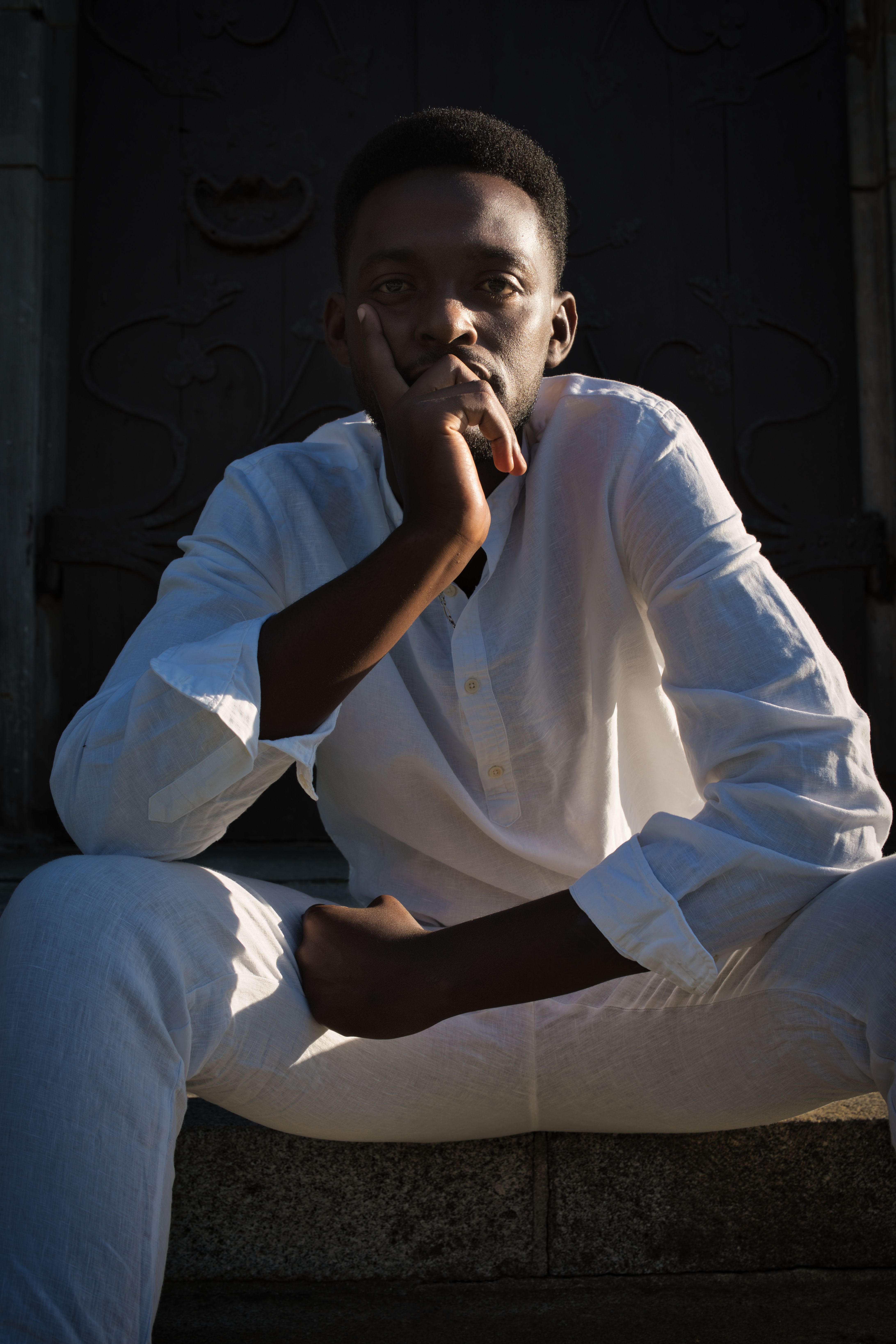 Contemplative man seated in white clothing, outdoors with moody lighting, Berlin.