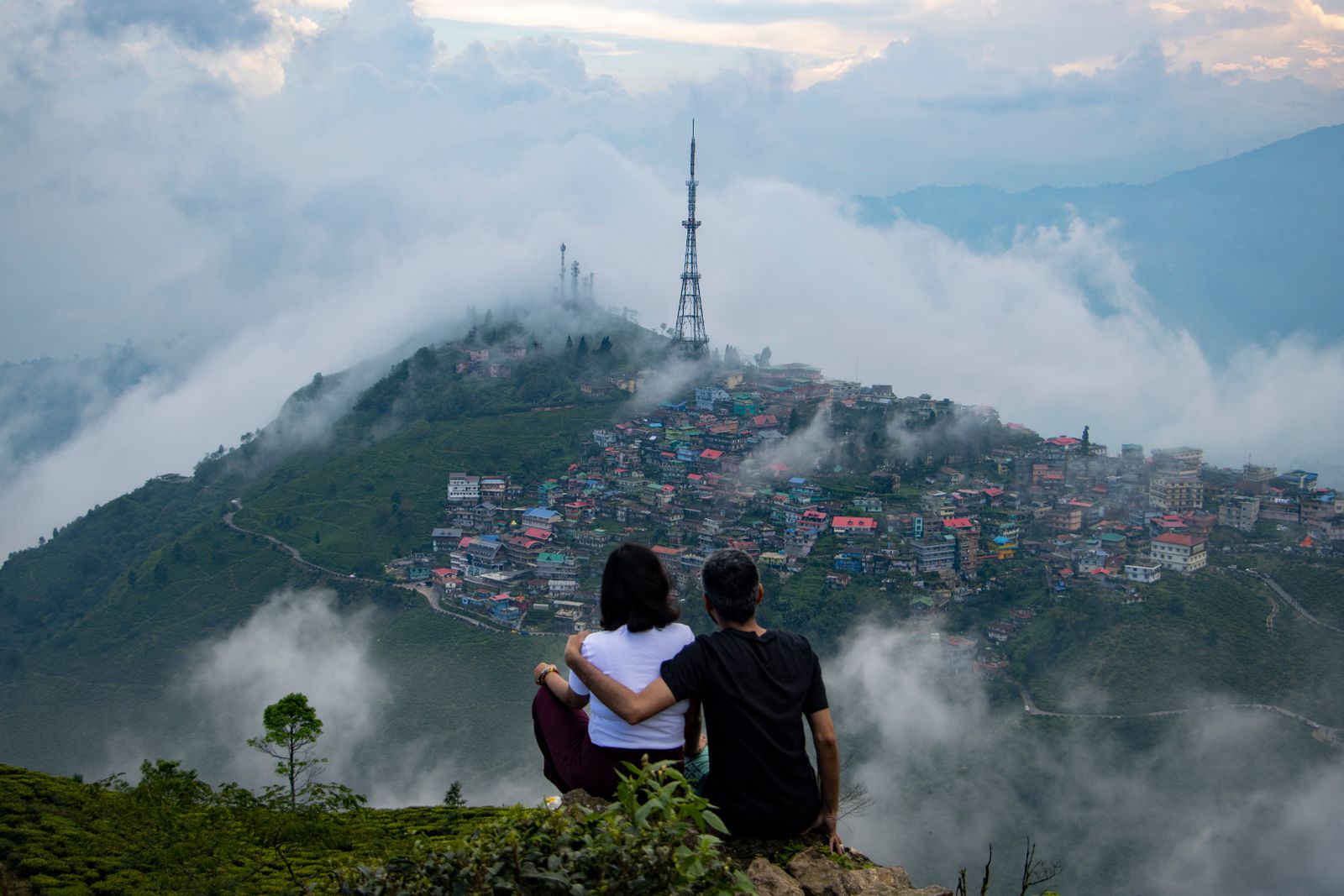 A couple sitting close on a tea garden ledge above Kurseong, watching cloud pour over the ridge town and its tall radio mast