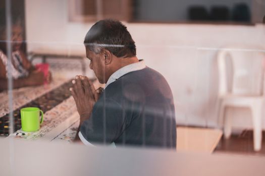 A man praying indoors at a table with a warm, comforting atmosphere.