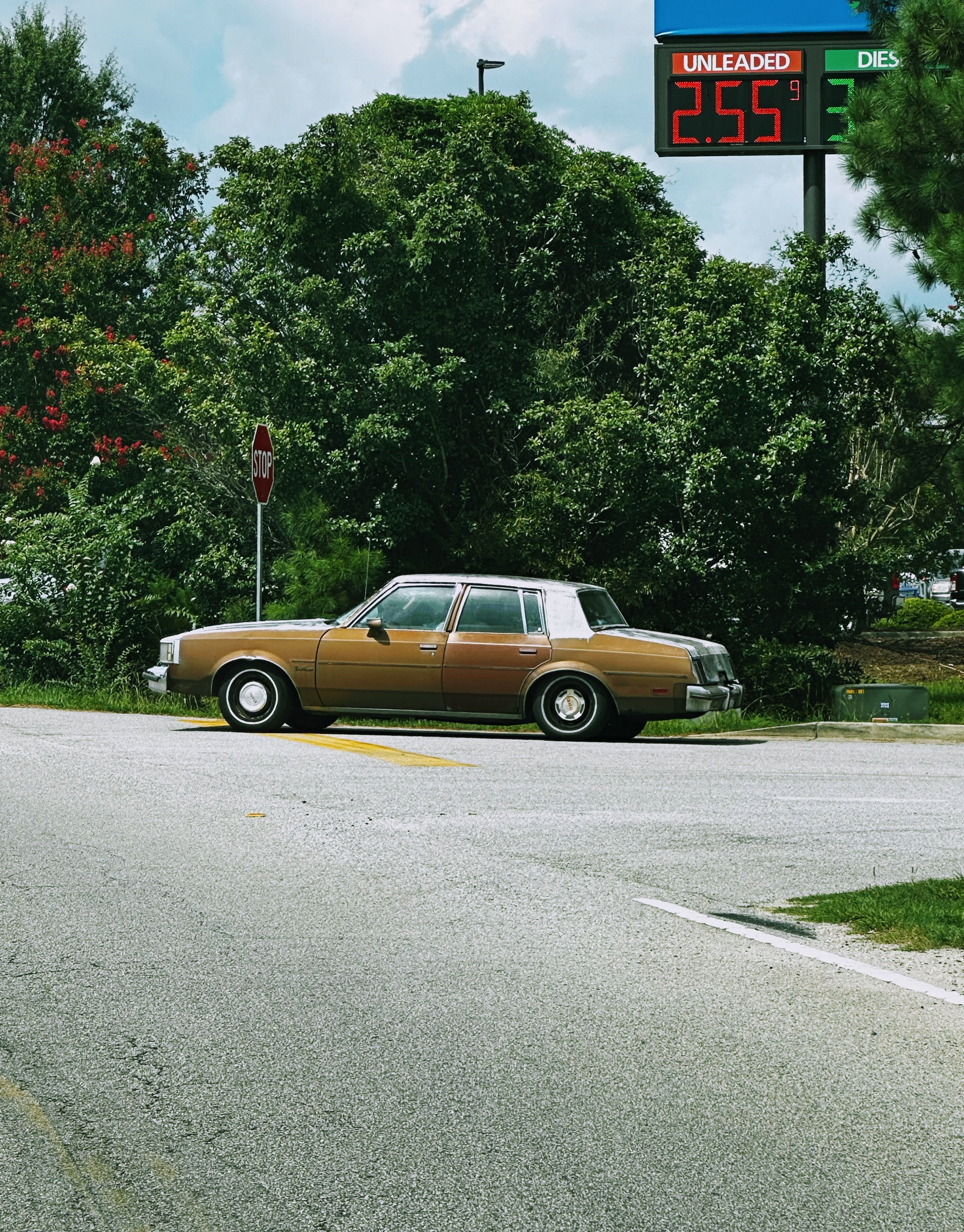 Brown vintage car parked near gas price sign in a green suburban area.