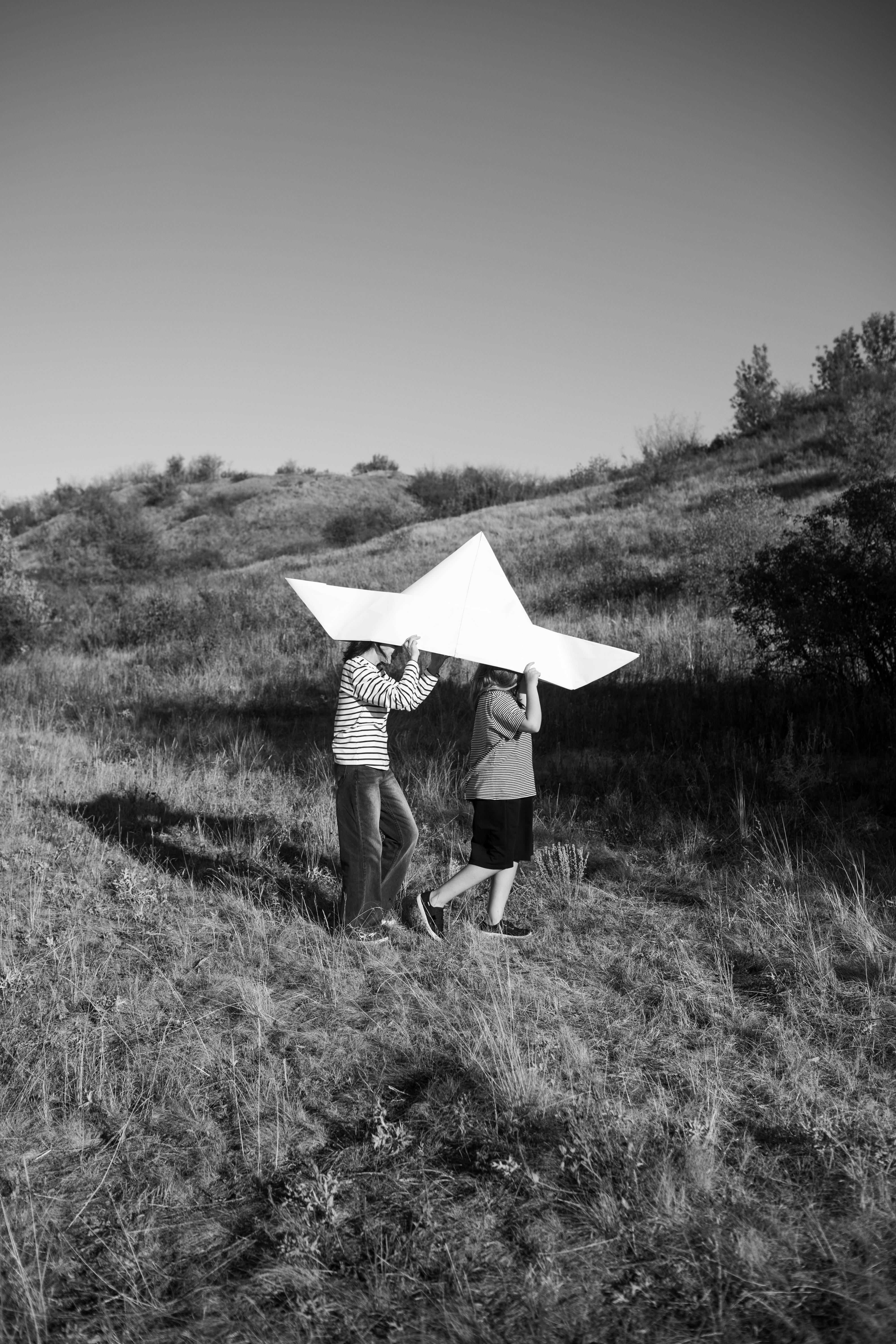Two children carry a giant paper airplane over a grassy hill, enjoying a sunny day outside.