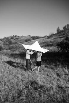 Two children carry a giant paper airplane over a grassy hill, enjoying a sunny day outside.