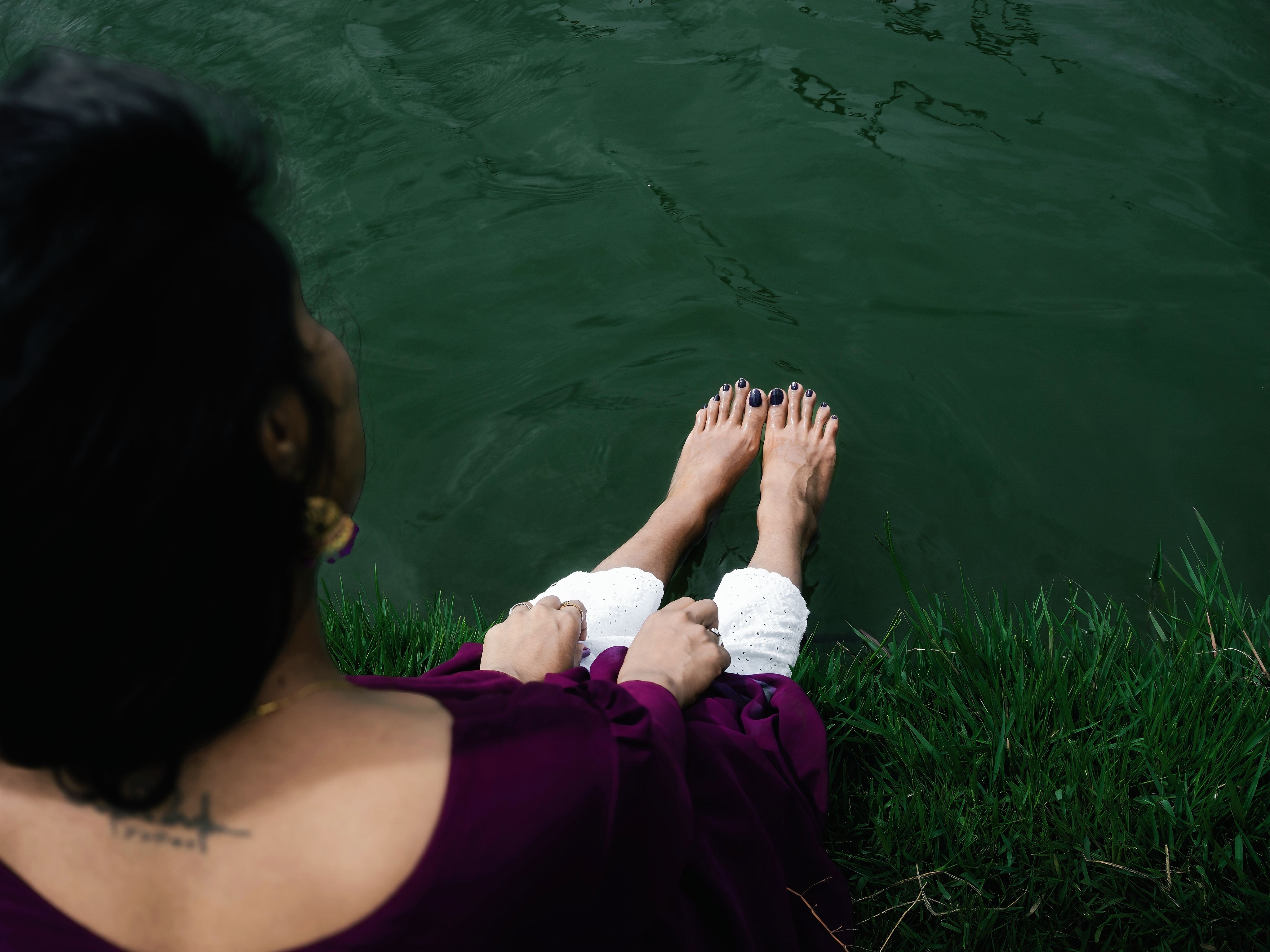 A woman sitting by a lake with her bare feet in the water, wearing a purple dress.