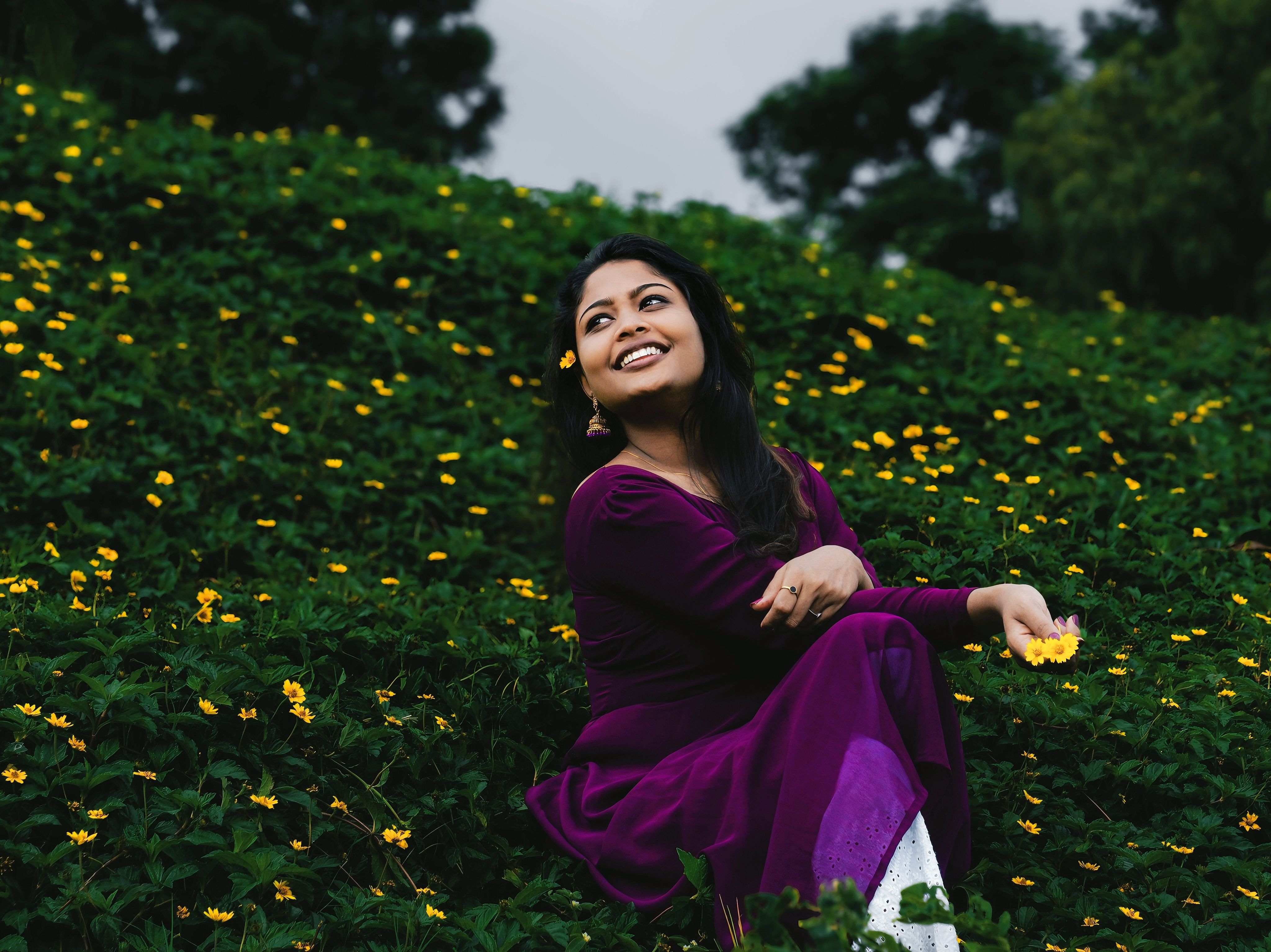 Joyful woman in purple dress sitting among vibrant yellow flowers outdoors.