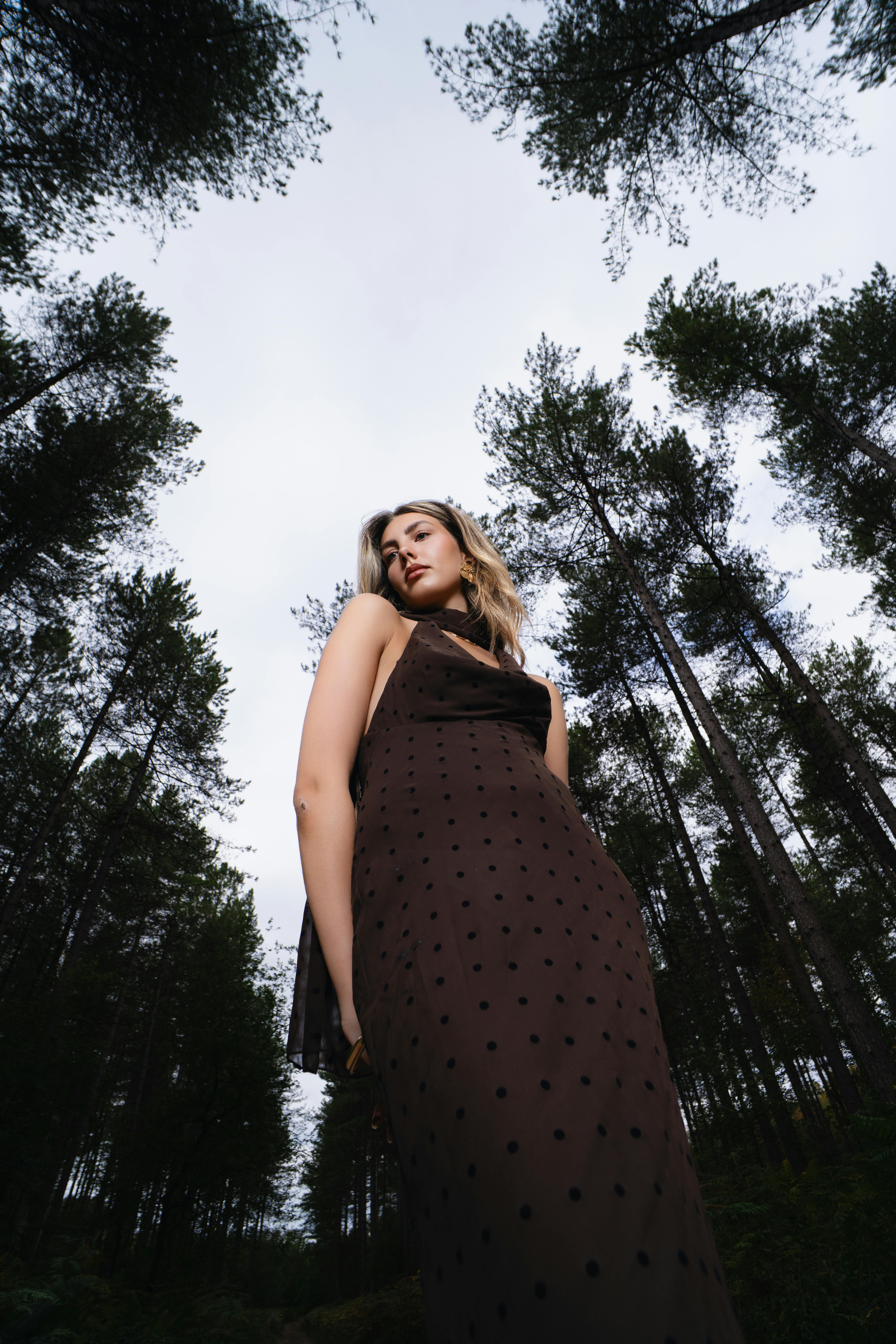 Model in stylish dress posing in a serene forest with tall trees.