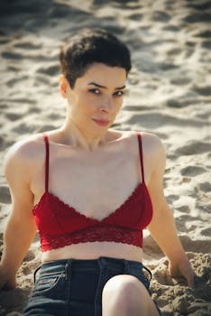 An adult woman in a red top enjoys a sunny day on the beach, leaning back on the sand.