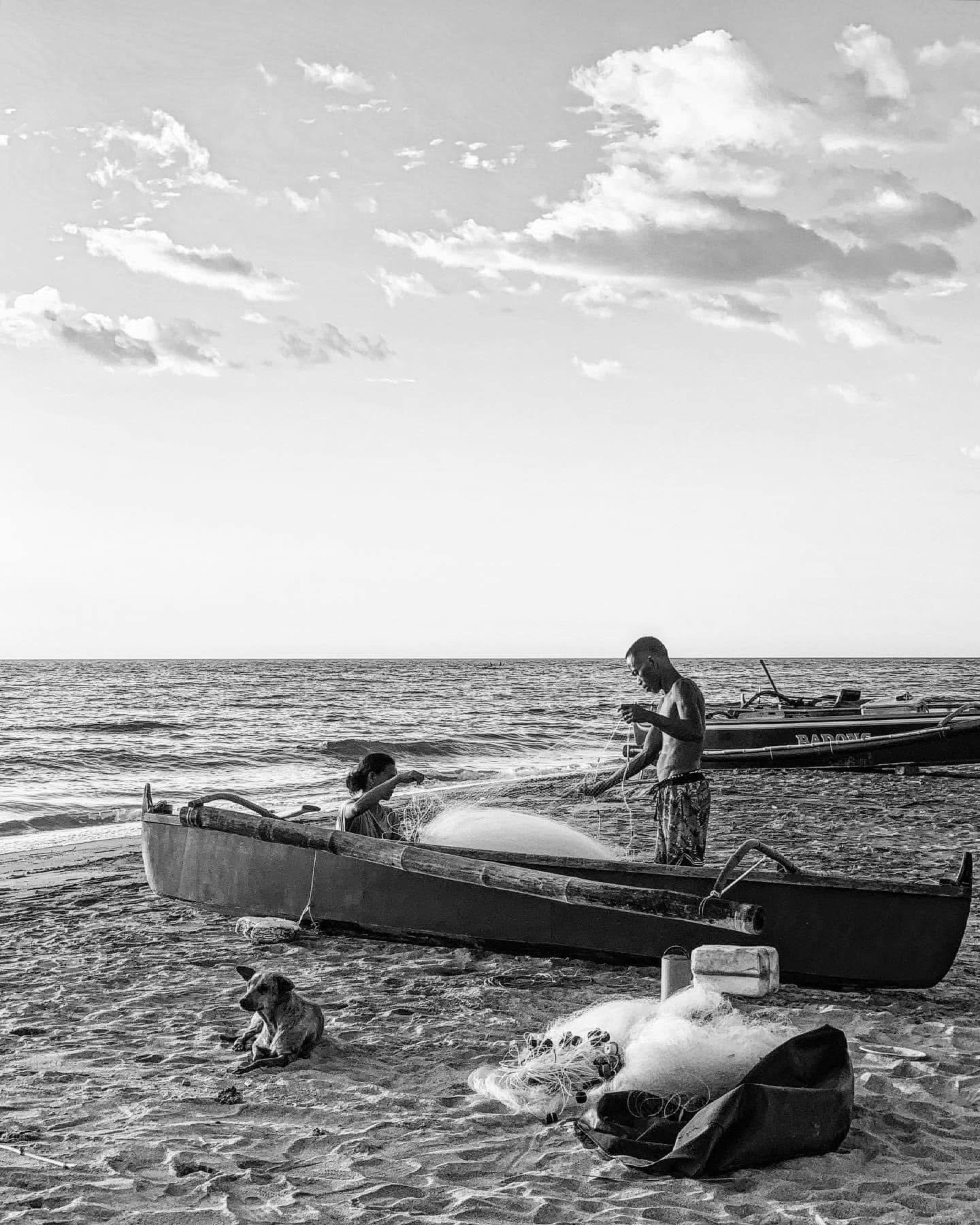 Two fishermen preparing nets by the sea with traditional boats in black and white.