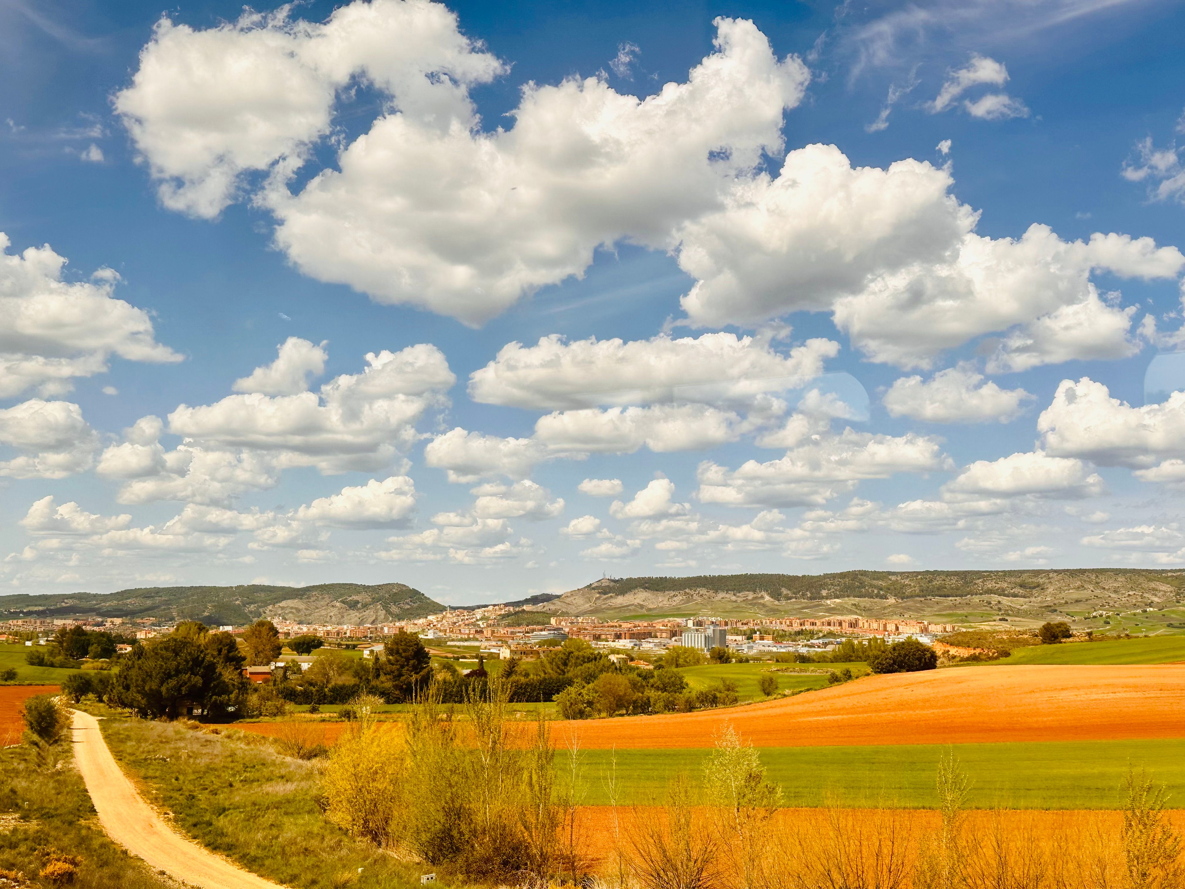 Beautiful countryside scene with lush fields, a dirt road, and rolling clouds under clear blue sky.
