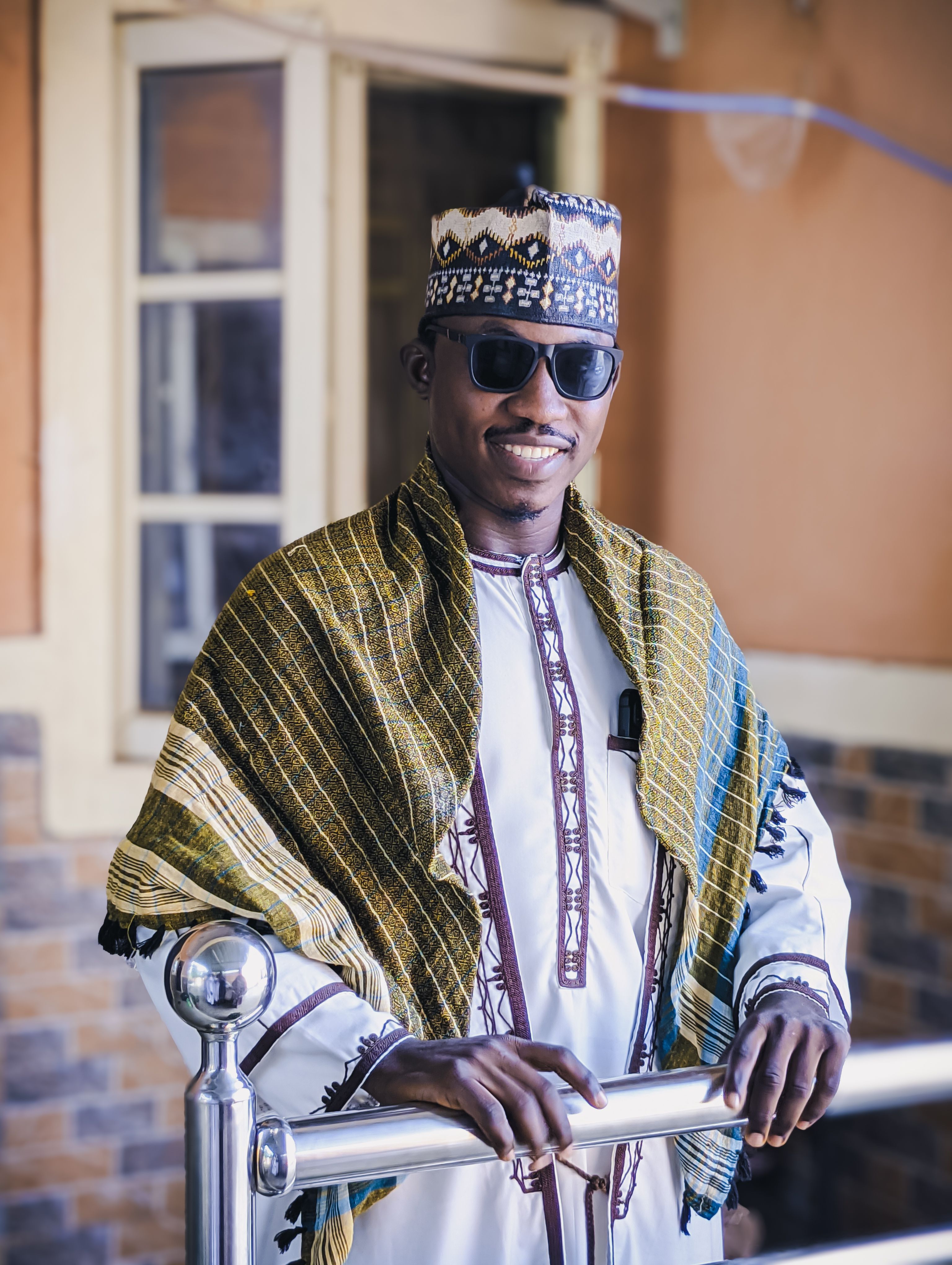 A confident man wearing traditional Nigerian attire and sunglasses, smiling indoors.