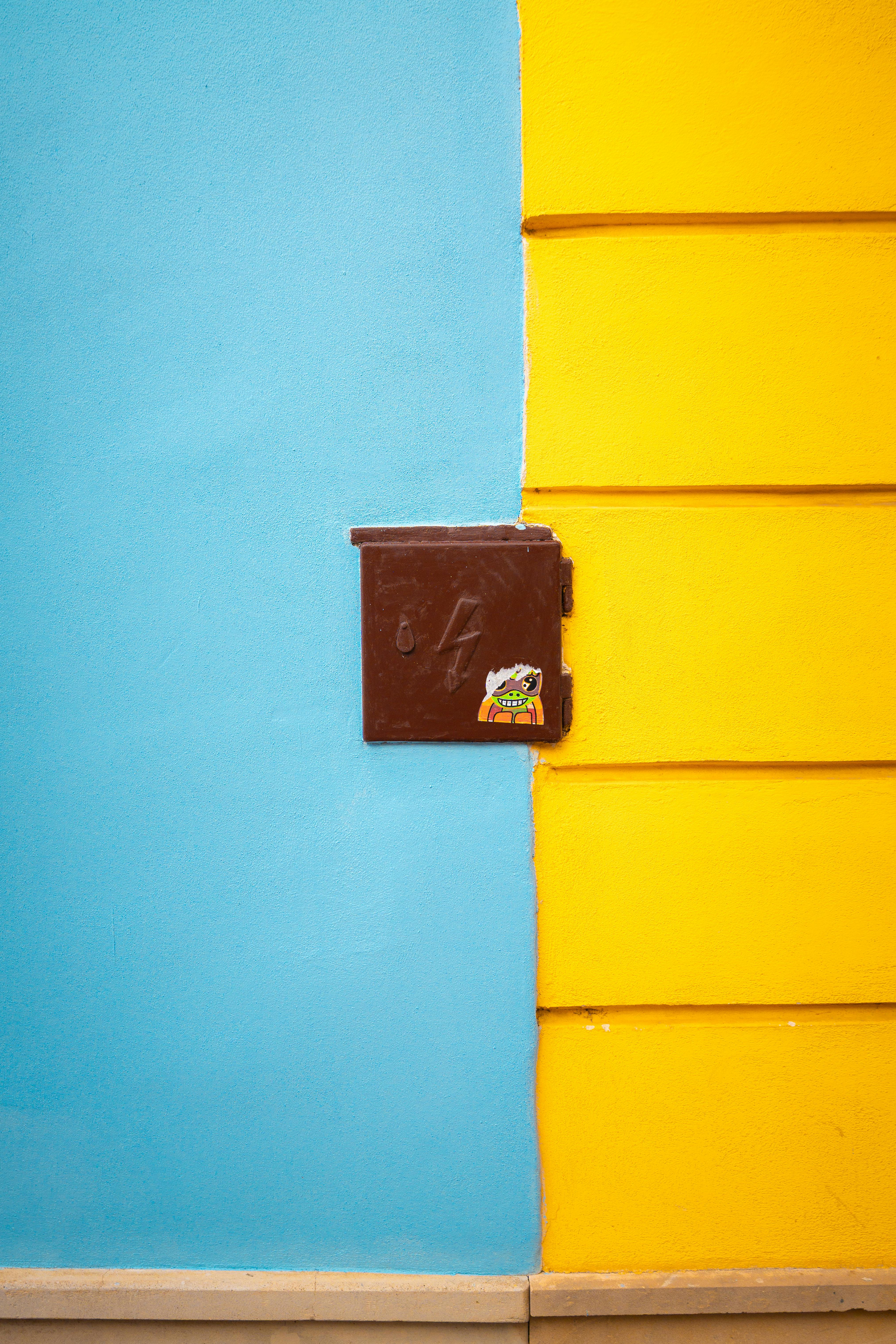 Kostenlos Eine leuchtend blaue und gelbe Wand mit einer zentralen braunen Box, die einen starken Kontrast erzeugt. Stock-Foto