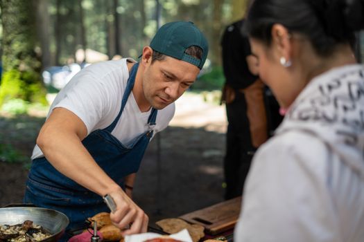 Chef cooking at an outdoor gathering, engaging with attendees in a lush forest setting.
