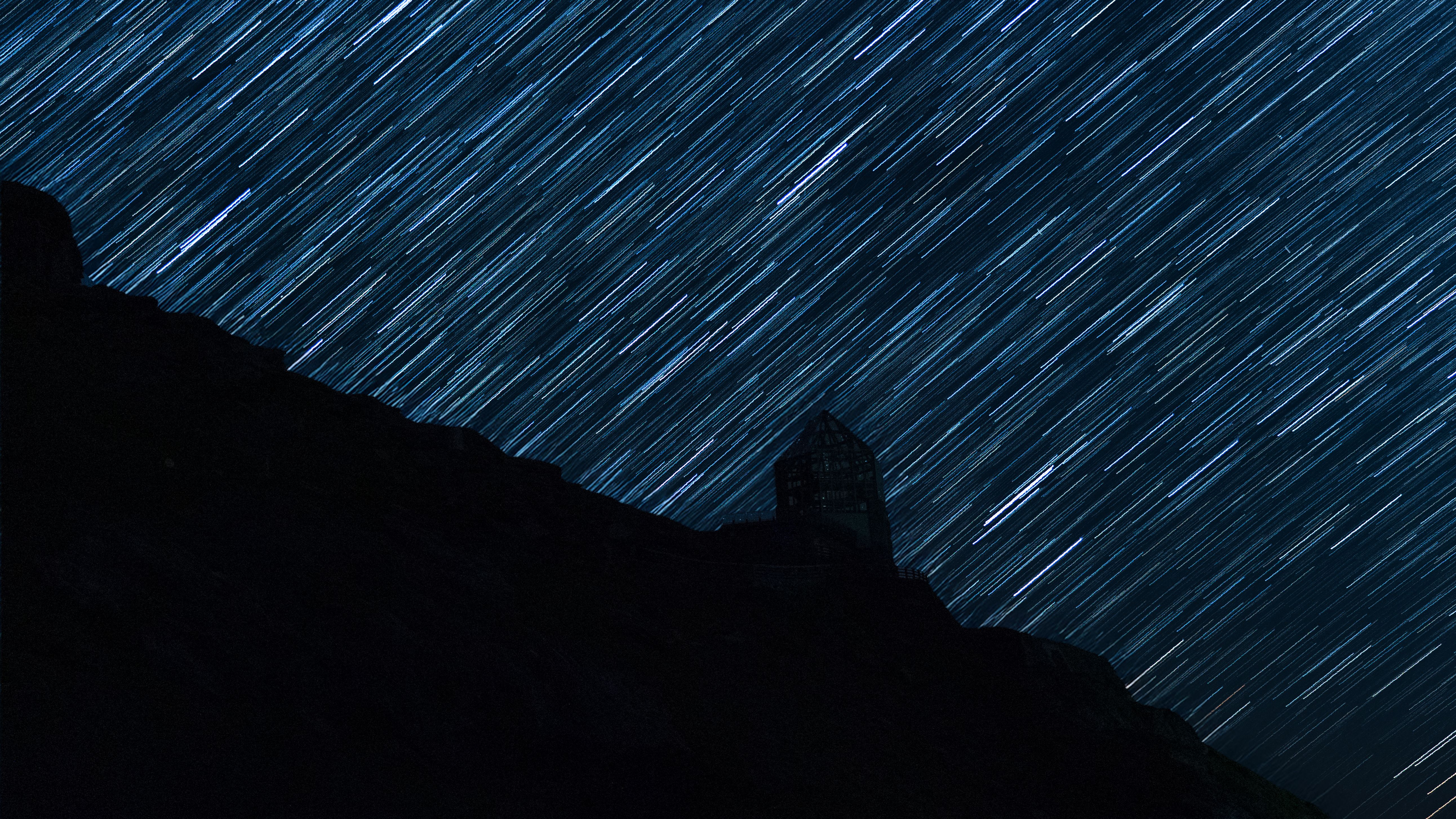 Long-exposure star trails creating a cosmic pattern above a mountain silhouette and observatory.