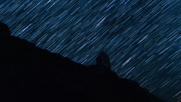 Long-exposure star trails creating a cosmic pattern above a mountain silhouette and observatory.