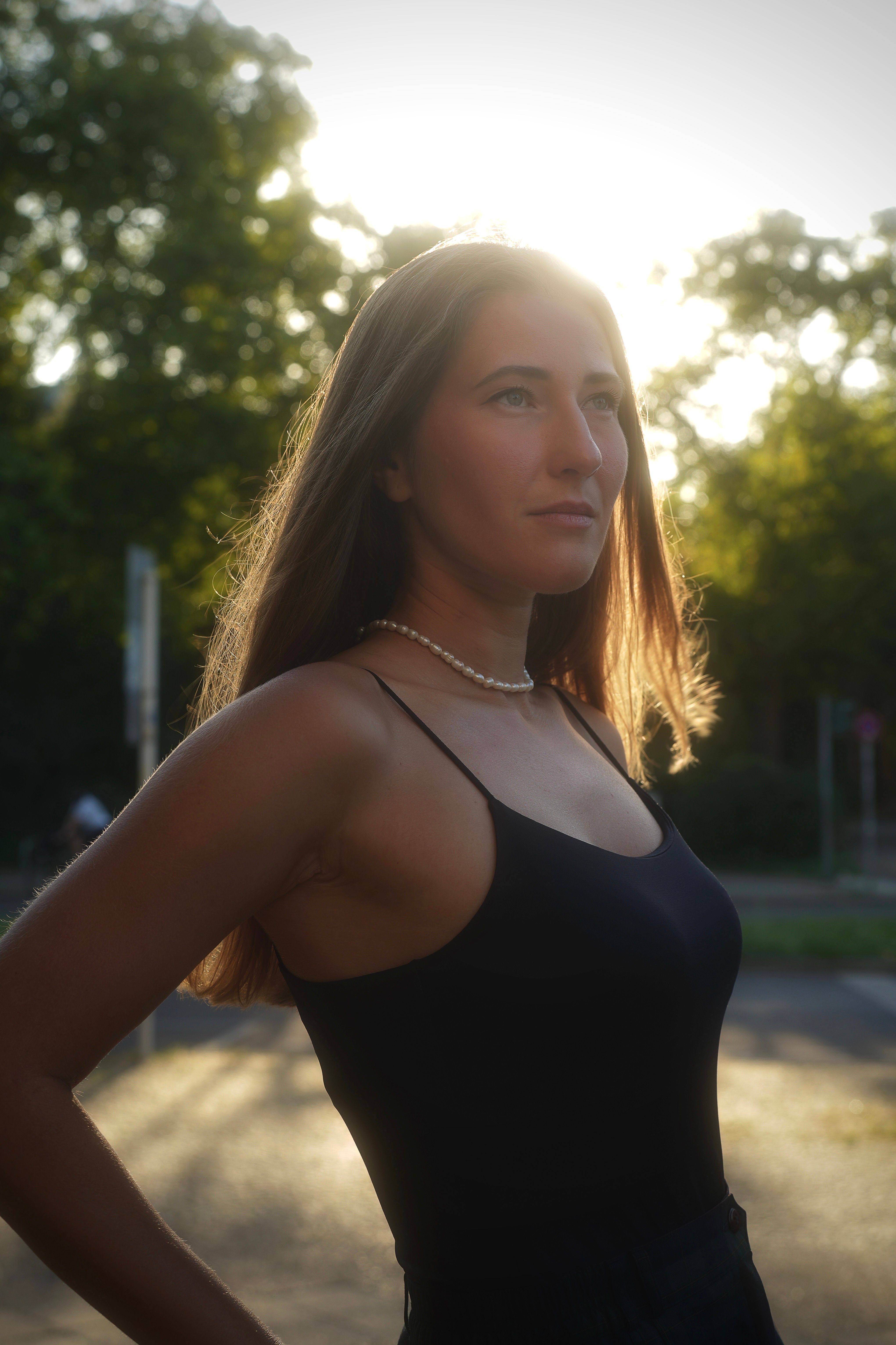 A young woman in a black dress standing confidently outdoors at sunset with trees in the background.