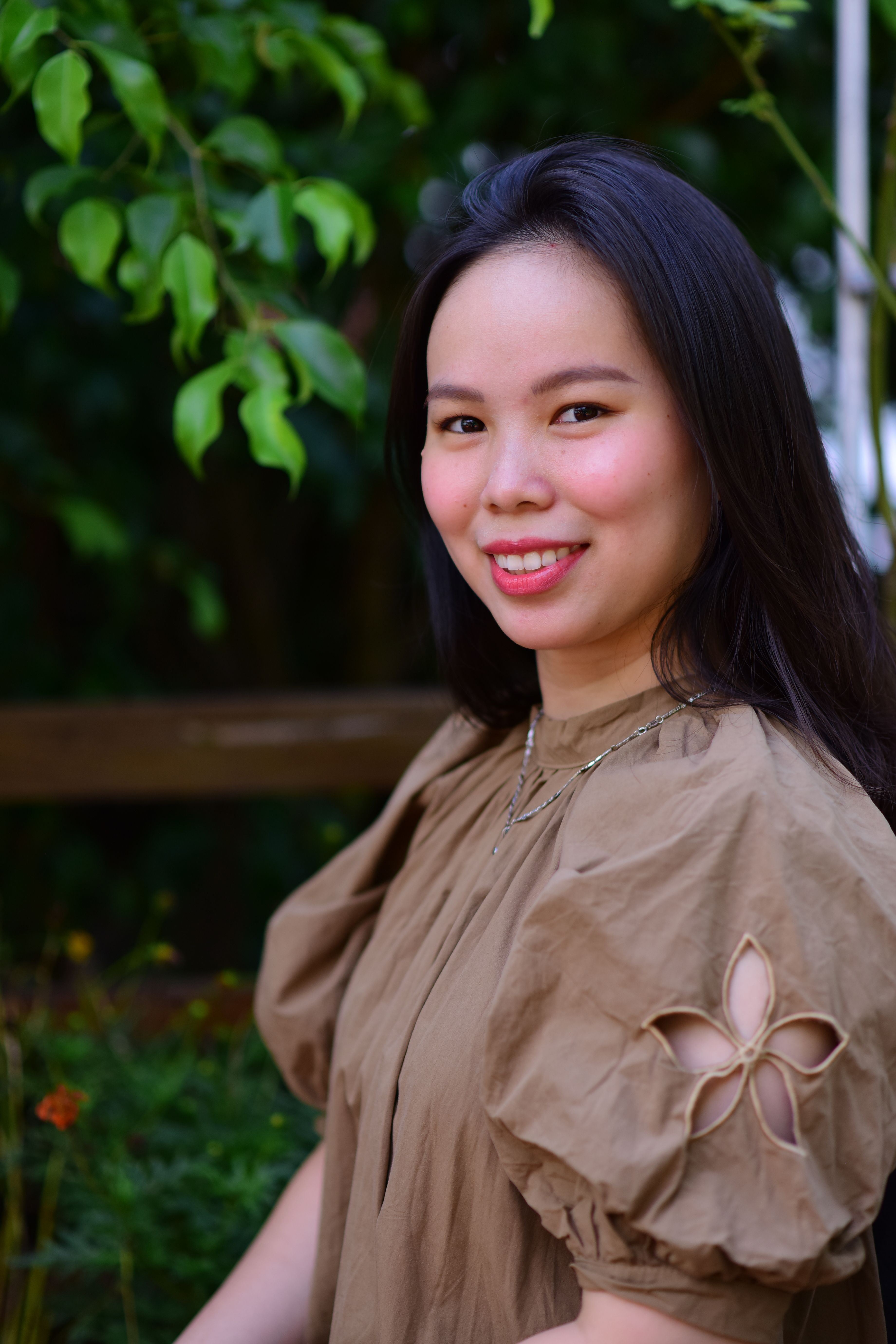 Smiling woman in a brown dress outdoors with green foliage background.