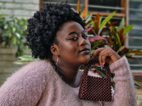 Free Confident woman with curly hair holding a chic beaded handbag in a colorful garden setting. Stock Photo