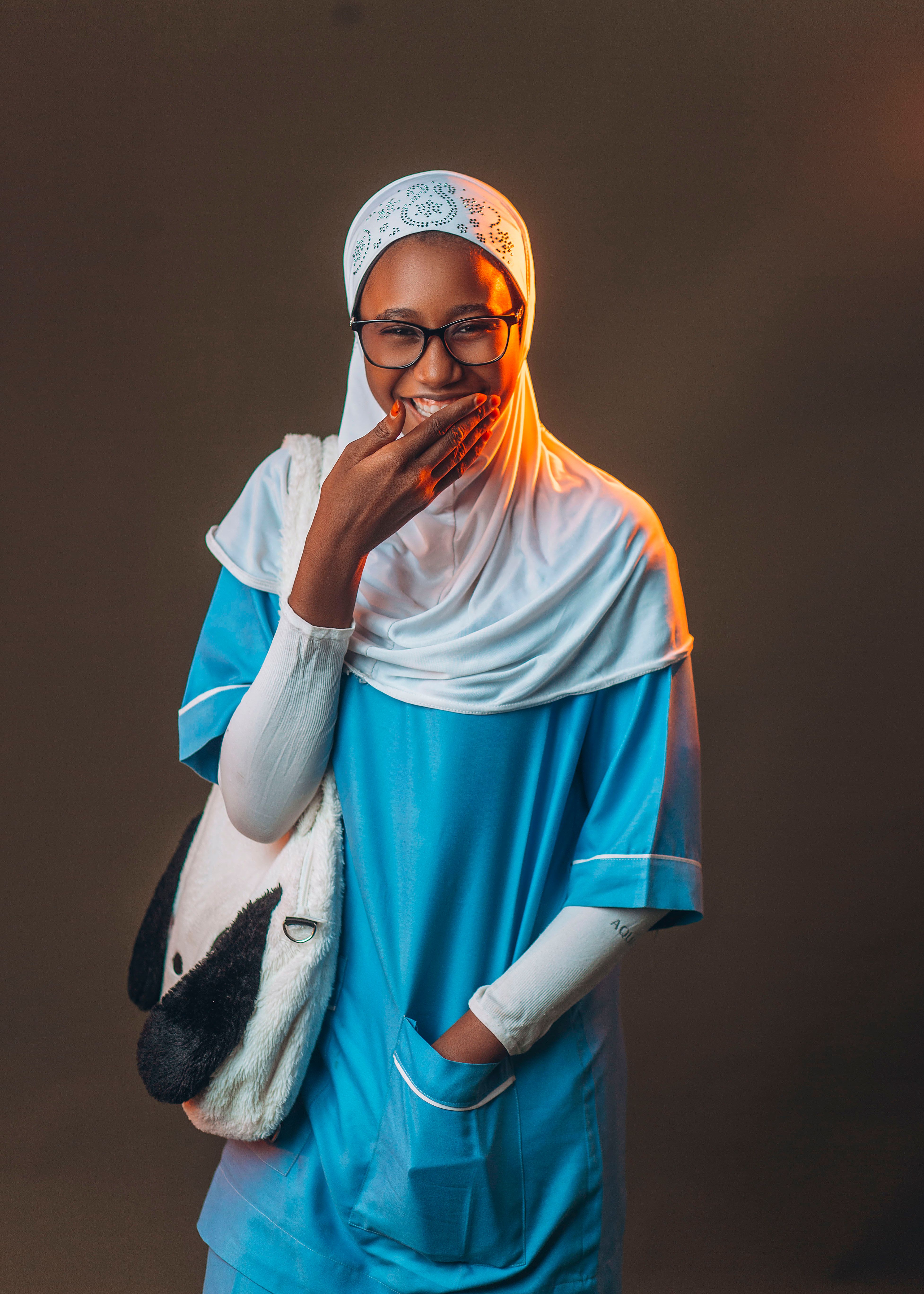 Smiling woman in blue attire with hijab holding a plush bag in a studio setting.