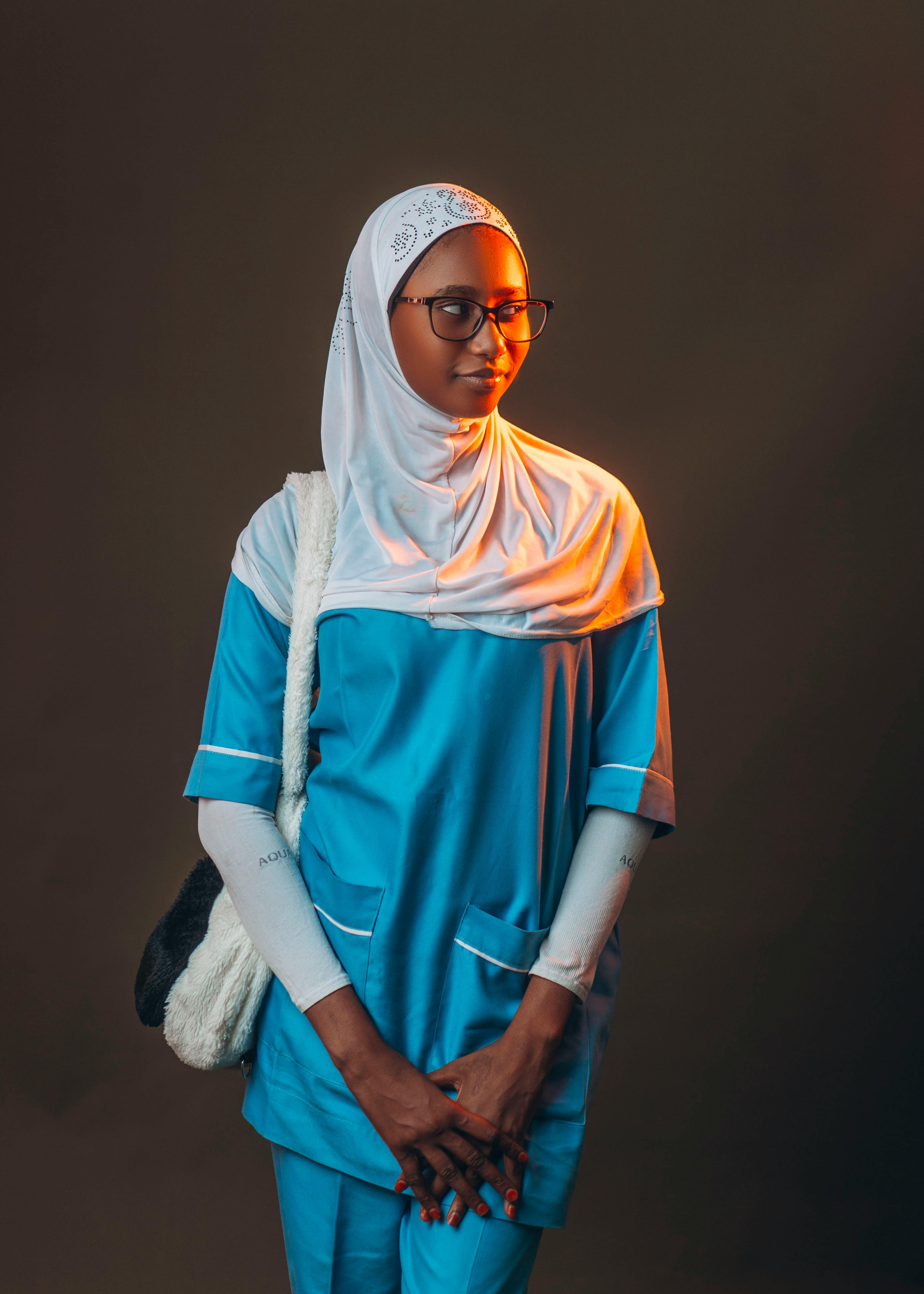 Confident female nurse in blue uniform and hijab standing with a warm light background.