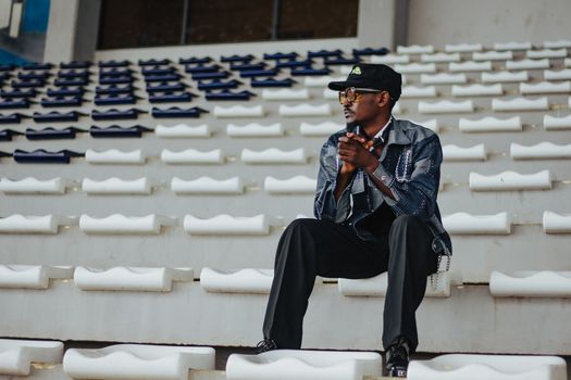 Man wearing denim and cap seated solo in a stadium, exuding a stylish and thoughtful vibe.