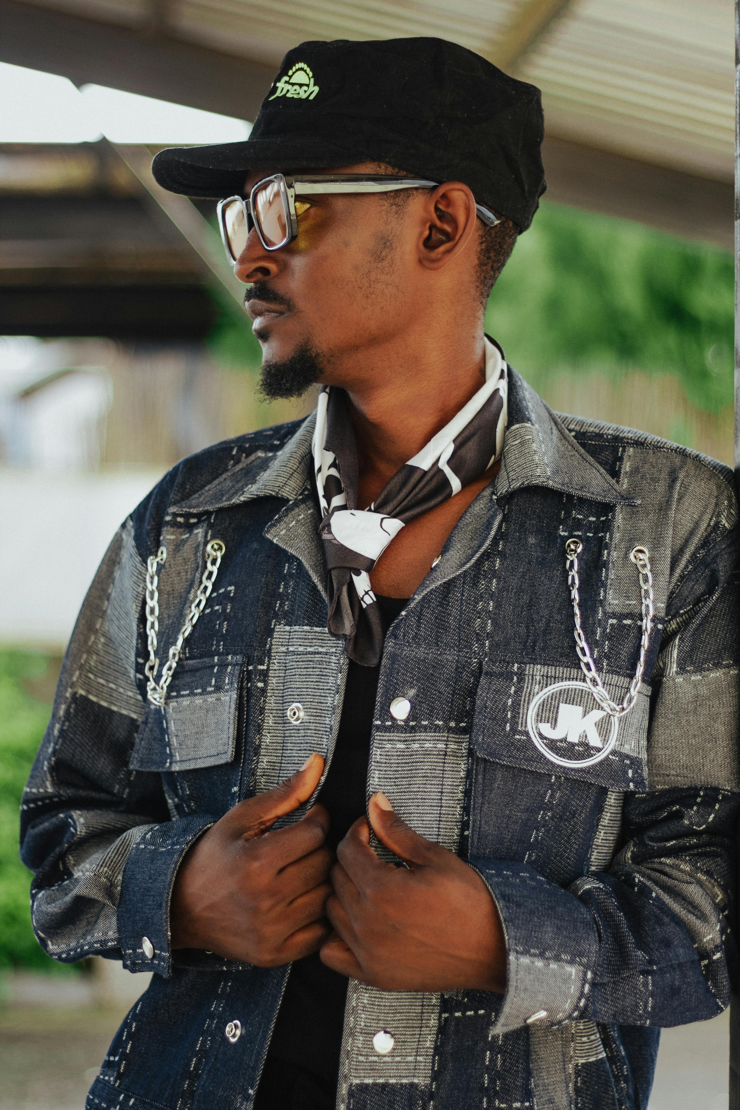 Young man in a patchwork denim jacket and cap standing outdoors, showcasing urban style.
